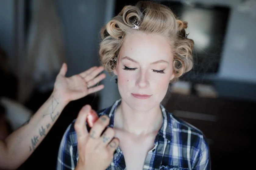 A woman with vintage styled blonde hair in curlers, eyes closed, getting her makeup done with a makeup brush, wearing a blue plaid shirt, in a room with blurred background.