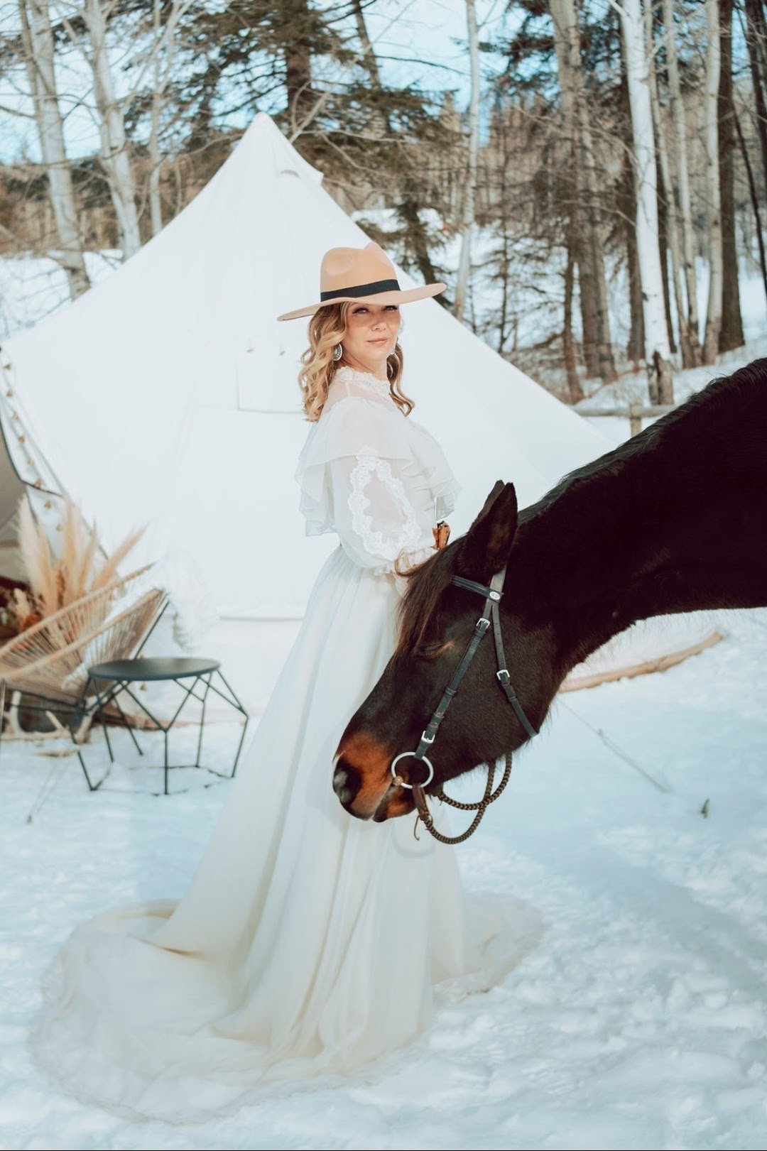 Woman in a white dress and wide-brimmed hat standing outdoors in a snowy forest, holding the reins of a brown and black horse.