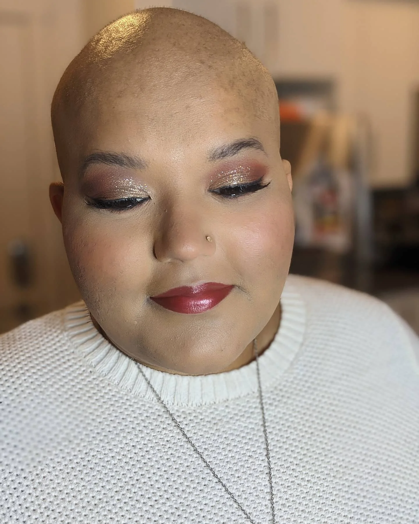 Close-up of a woman with a shaved head, wearing makeup with glitter eyeshadow and red lipstick, and a white sweater.