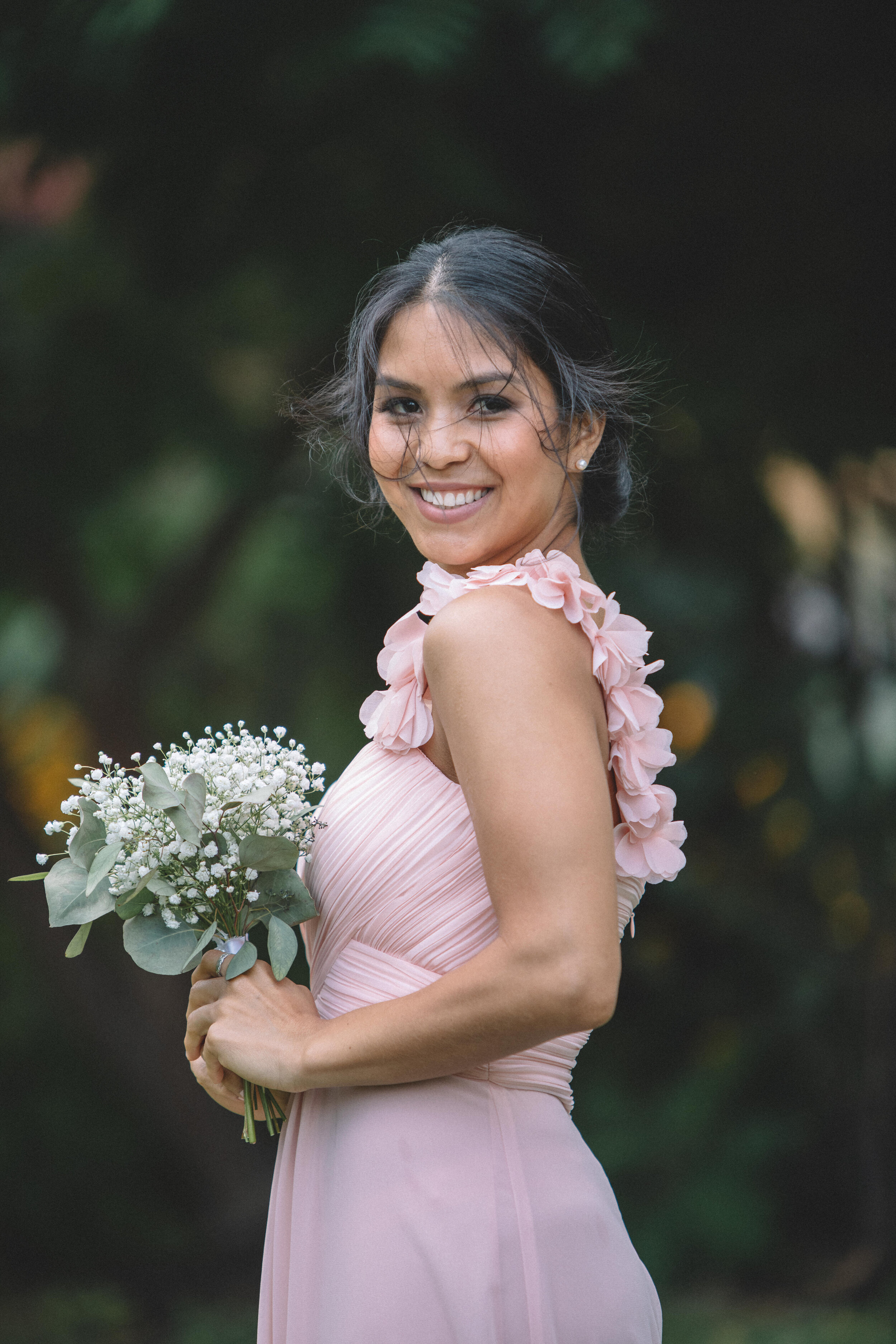 A woman in a pink dress with floral embellishments on the shoulders, smiling, holding a bouquet of white flowers and greenery, standing outdoors with dark blurred background.