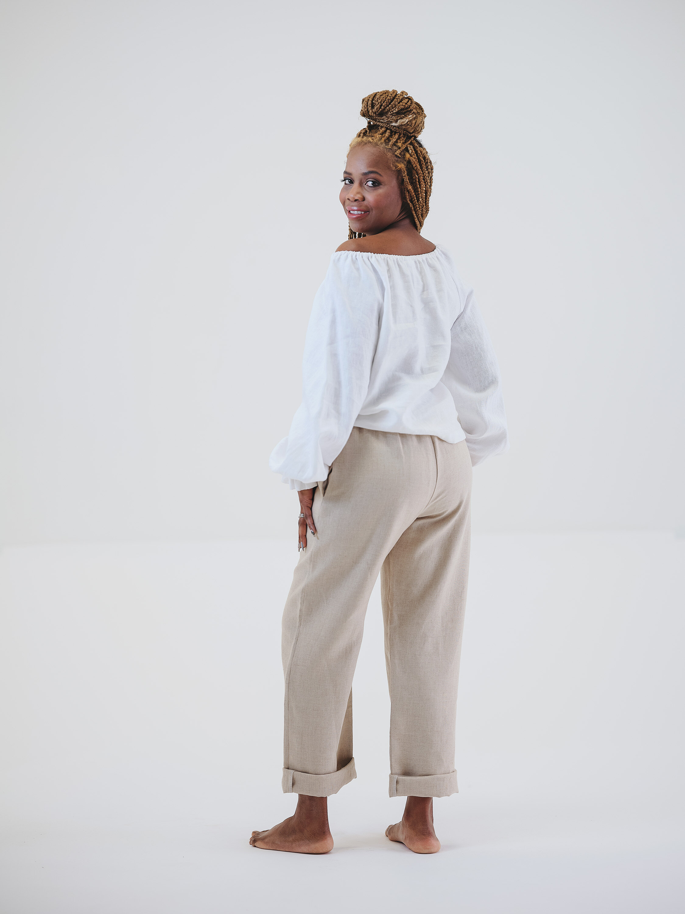 A woman with light brown skin and blonde braids styled in a topknot, wearing a white off-shoulder blouse and beige pants that are rolled up at the cuffs, standing barefoot against a plain white background, looking over her shoulder with a slight smil