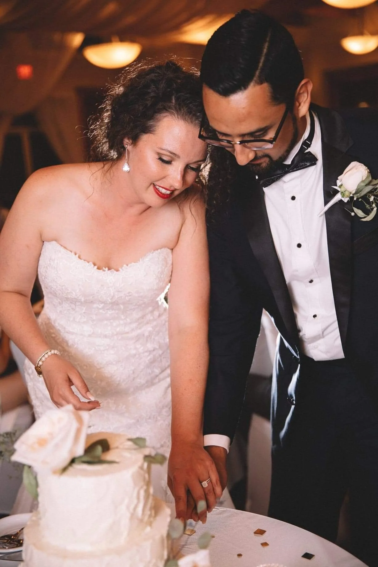 A bride and groom cutting their wedding cake together at a reception. The bride wears a strapless white dress with lace details, and the groom is in a black tuxedo with a bow tie and boutonniere. They are smiling and looking down at the cake.
