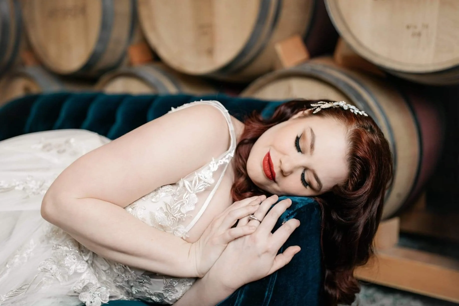 A woman with red hair and makeup, wearing a white lace dress and a hair accessory, peacefully sleeping while lying on a dark blue velvet surface with wooden barrels in the background.