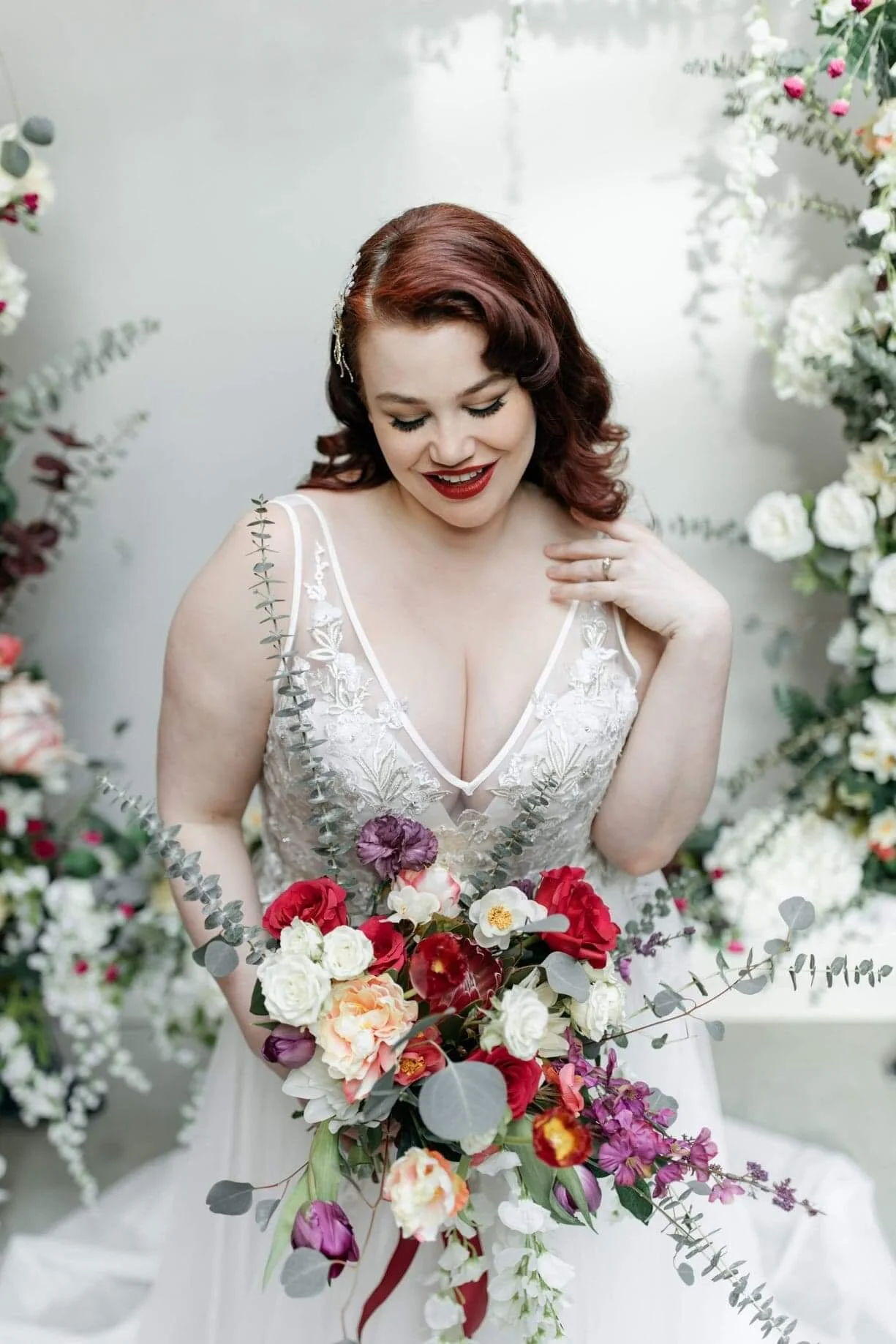 A woman with red hair in a vintage style, wearing a white wedding dress with lace details, holding a large bouquet of colorful flowers, and standing amidst floral decorations.