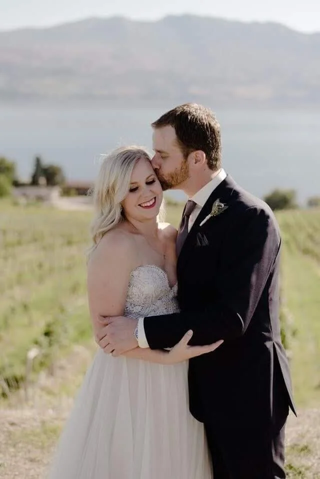 A bride and groom embrace outdoors during their wedding, with a lake and mountains in the background. The groom kisses the bride on the forehead, and they are smiling.