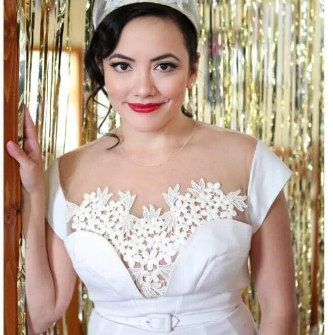 A woman wearing a white dress with floral lace detailing on the chest, standing in front of a gold tinsel backdrop.