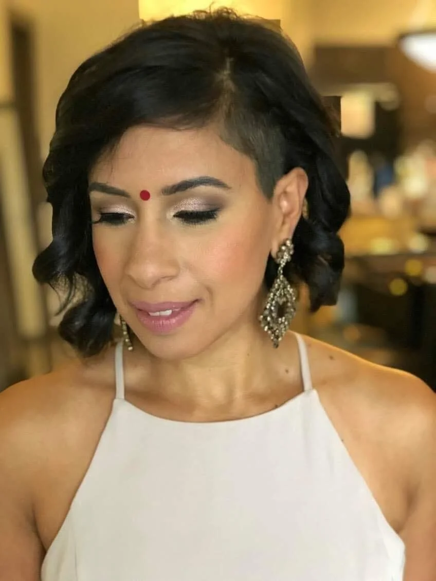 Close-up of a woman with short black hair, wearing a white sleeveless top, dark eye makeup, a red bindi on her forehead, and large dangling earrings, looking slightly downward with a gentle smile.