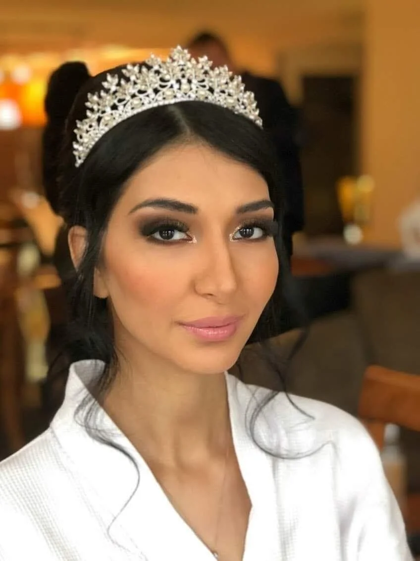 A woman wearing a tiara and white top, with dark hair styled with soft waves, makeup with dark eyeshadow and light lipstick, sitting in a warmly lit indoor setting.