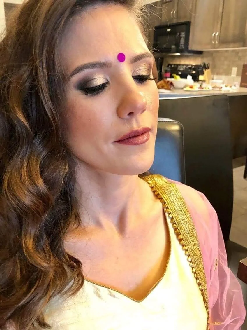 A woman with long brown wavy hair and makeup, wearing a pink and gold traditional Indian outfit, sitting in a kitchen.