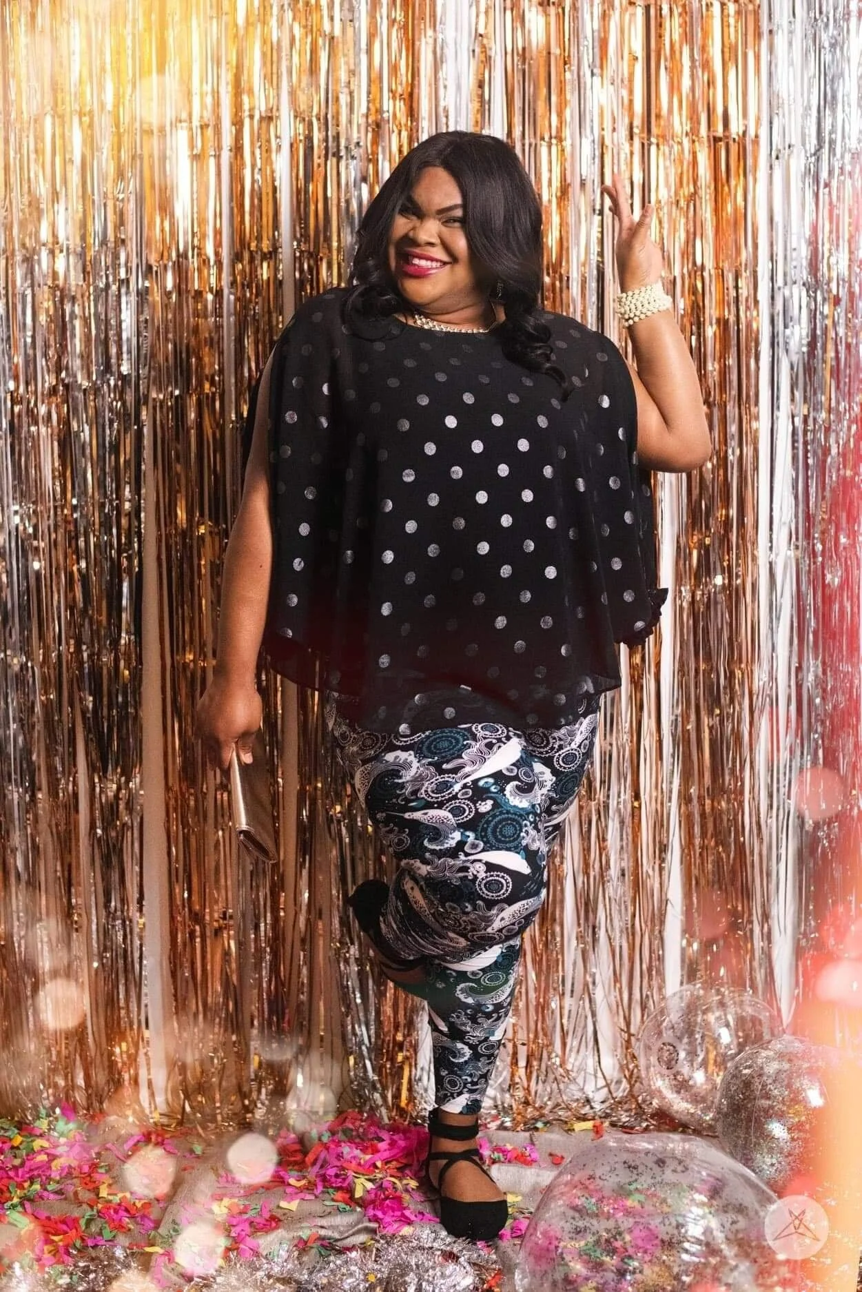 A woman in a black dress with white polka dots, black and white patterned pants, and black shoes is standing in front of a shiny, metallic gold curtain backdrop. She is smiling, winking, and holding a clutch purse, with colorful confetti, balloons, a