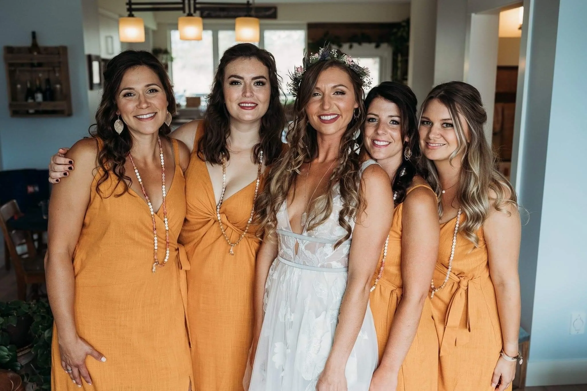 A group of five women dressed in summer attire, standing indoors, smiling at the camera. One woman in the center is wearing a white lace dress and a floral crown, suggesting a bride, while the others are wearing matching yellow dresses, indicating th