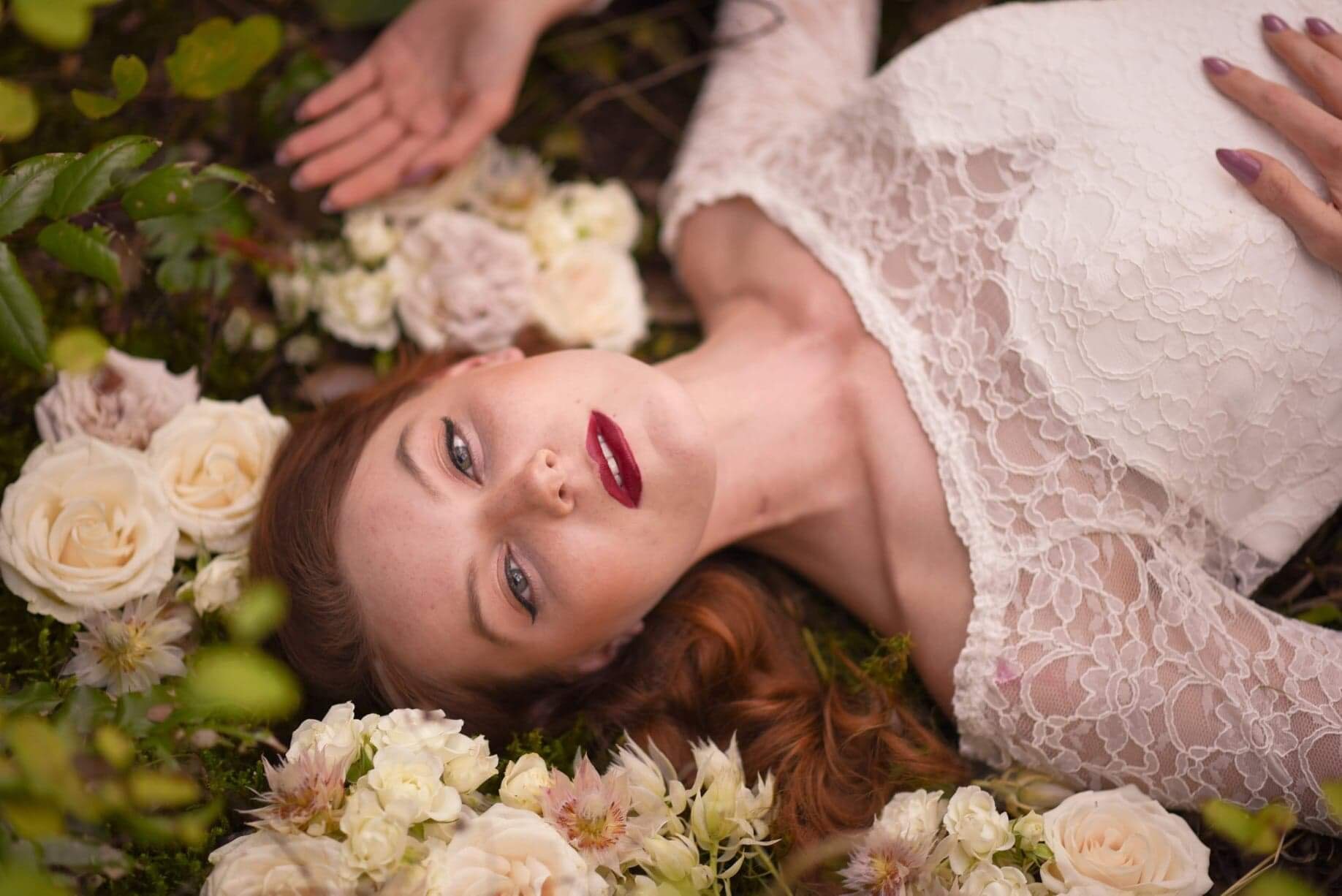 A woman with long red hair lying on the ground among white and pink roses and green foliage, wearing a white lace dress, with her eyes open and head tilted to the side.