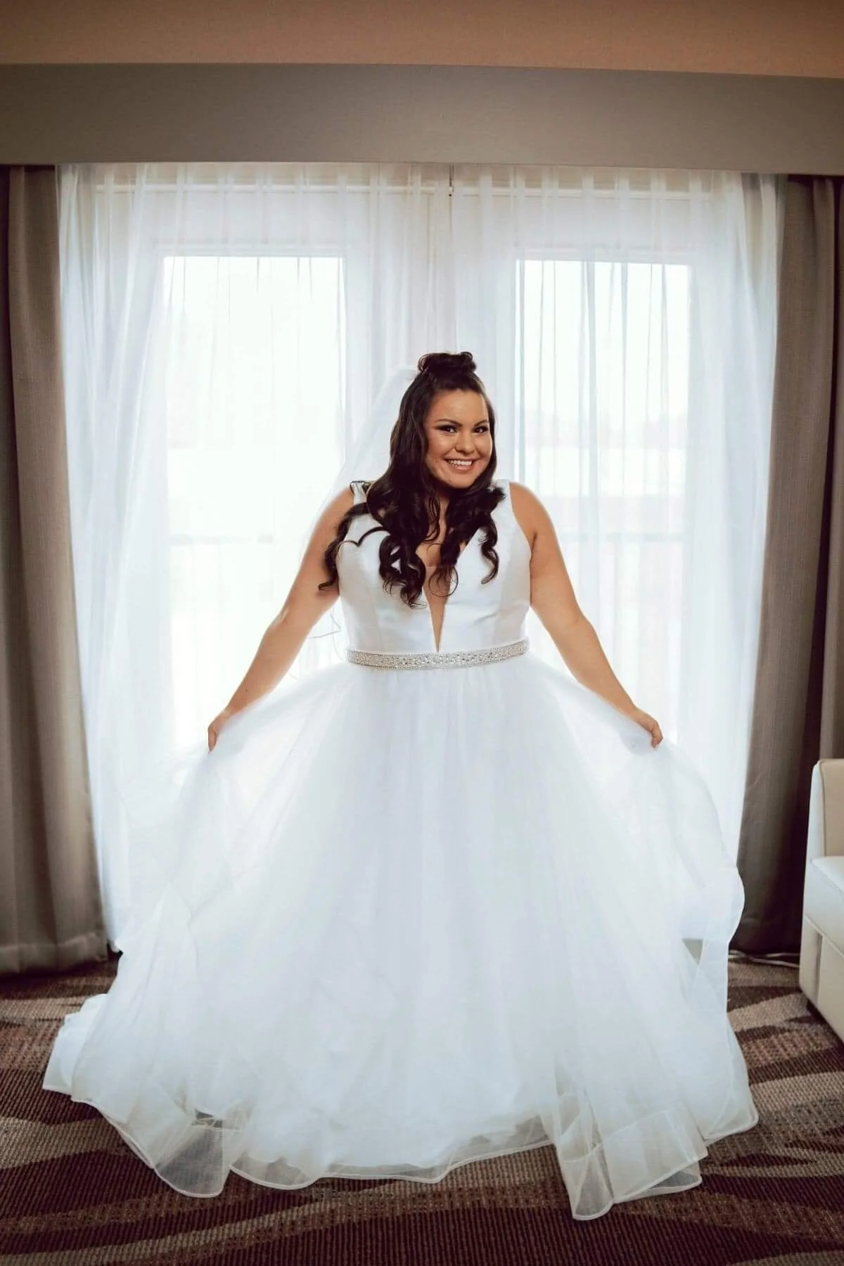 A smiling woman in a white wedding dress holding out the skirt, standing in front of a window with white curtains.
