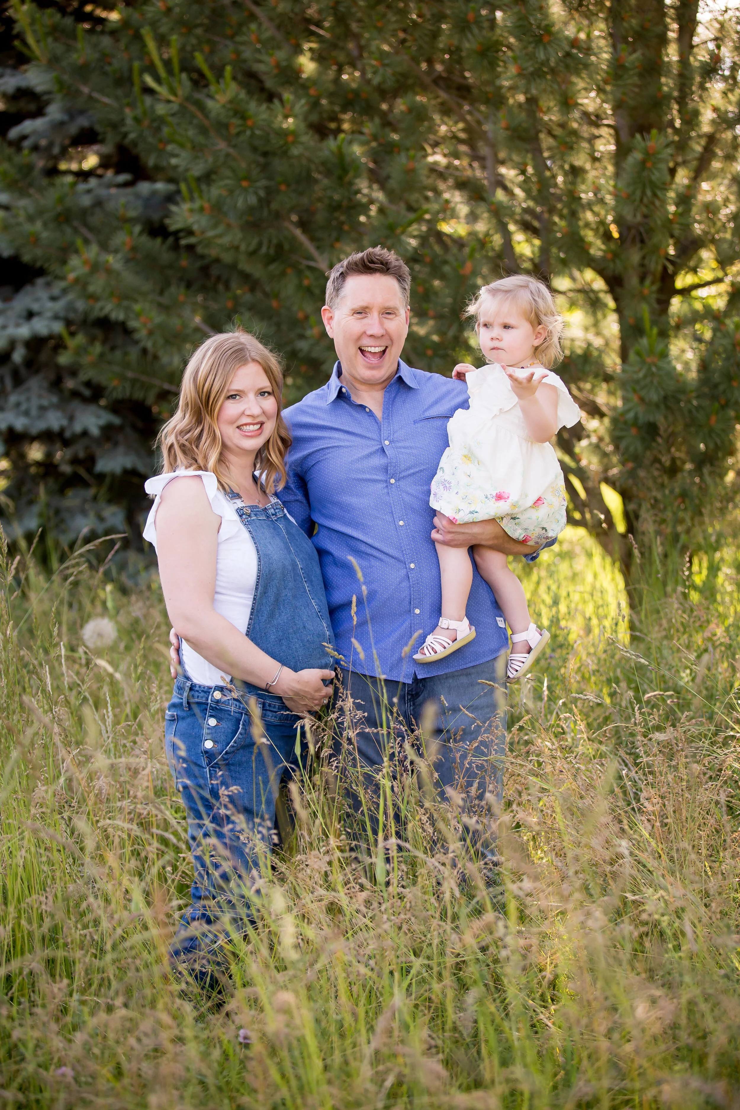 A family of three standing outdoors in a grassy field with trees in the background. The father is holding a young girl, and the mother stands next to him, all smiling.