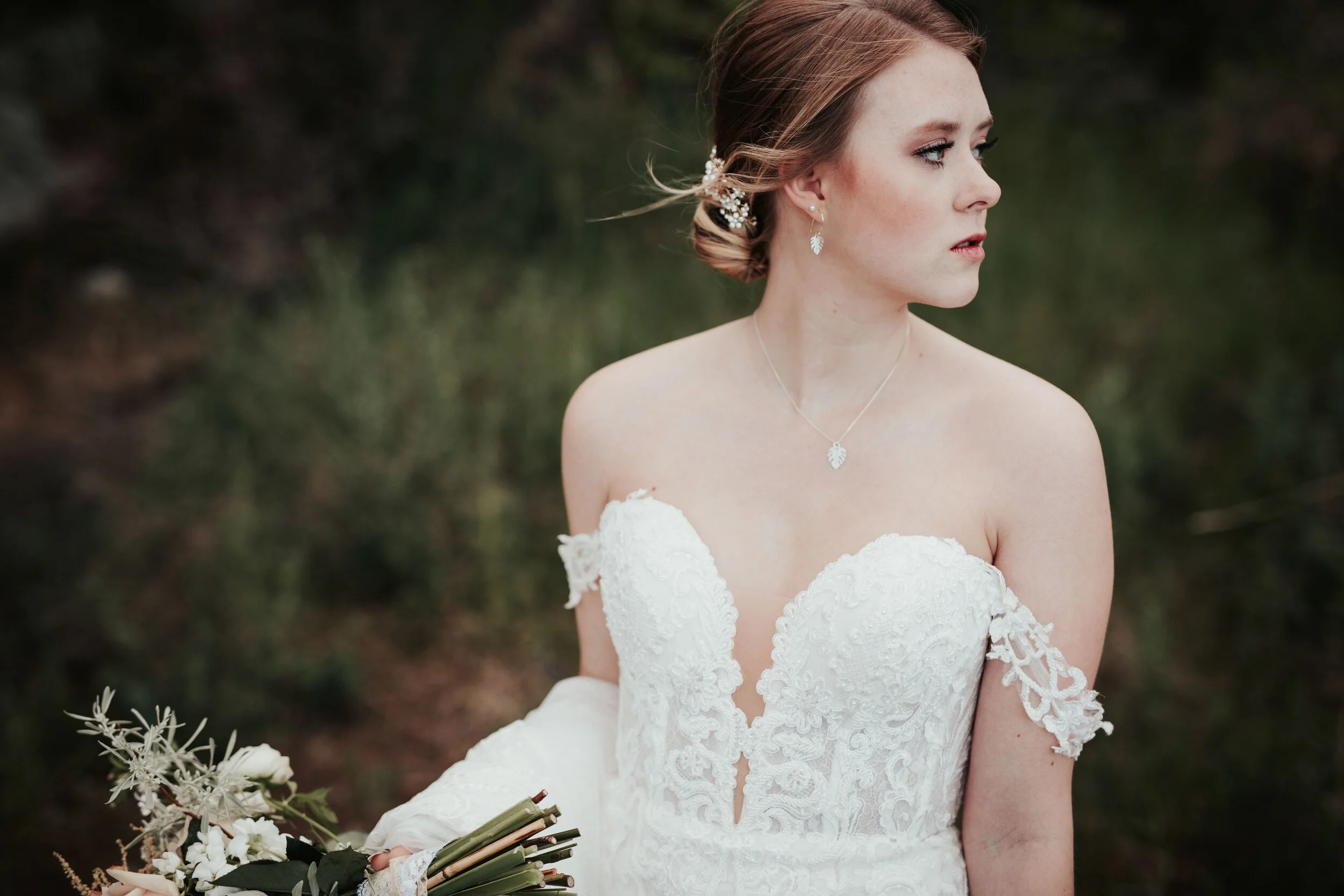 A bride wearing a lace wedding dress with off-shoulder sleeves, silver floral jewelry, and holding a bouquet of white flowers and greenery, sitting outdoors with a blurred green background.