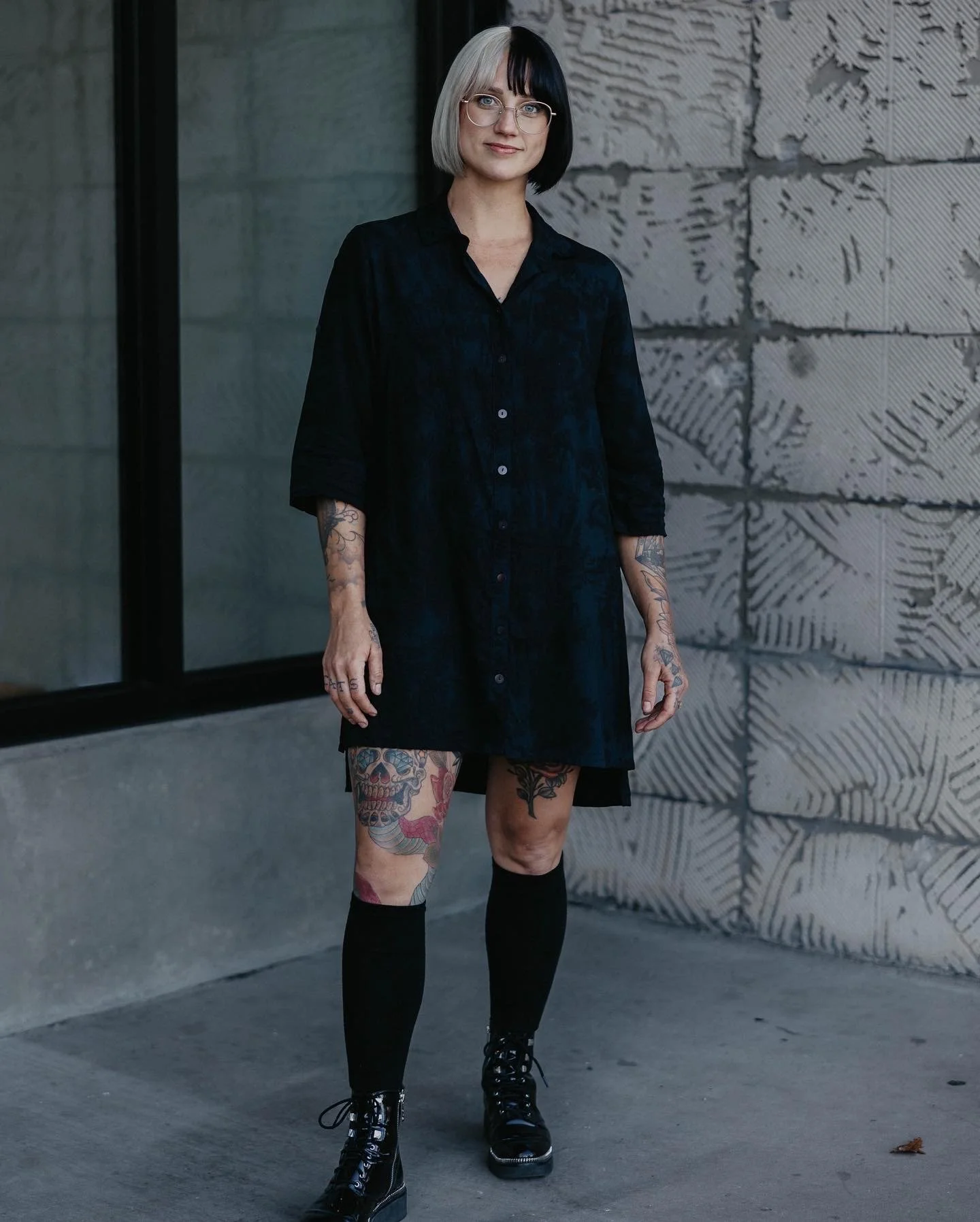 A woman with short black and platinum blonde hair, glasses, a black dress, black knee-high socks, and black boots, standing outside near a textured concrete wall and a window.