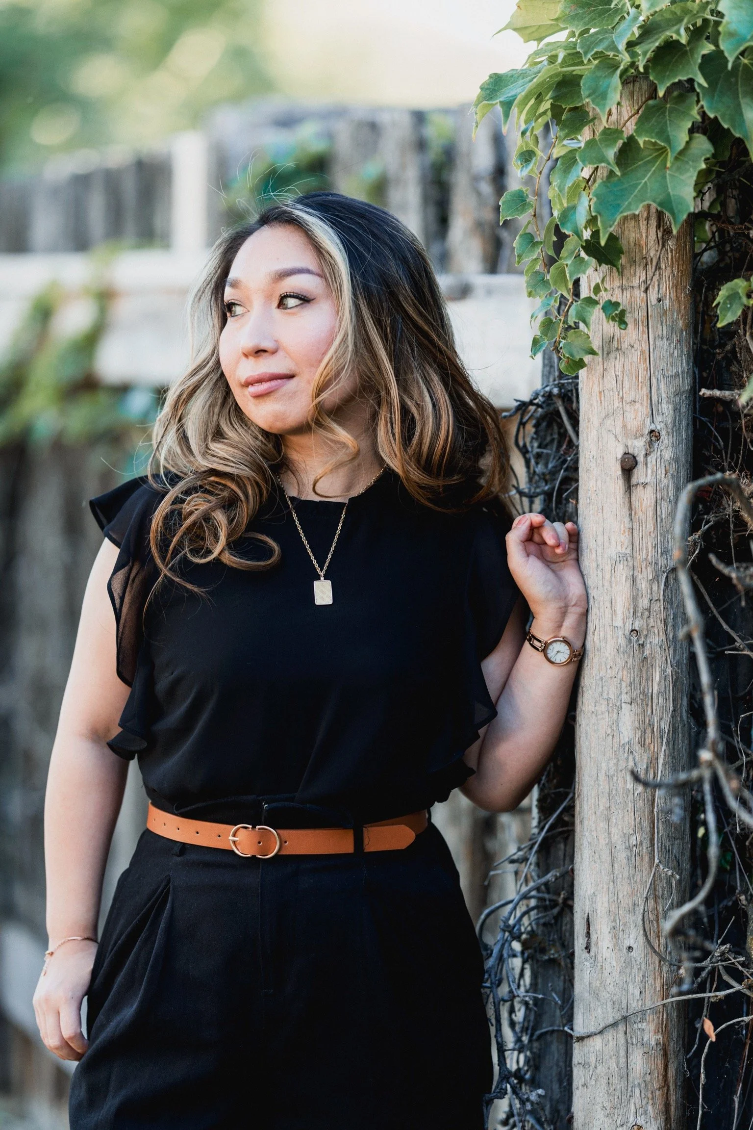 A woman with wavy hair and makeup wearing a black top with ruffled sleeves and a tan belt, standing outdoors against a wooden fence with ivy.