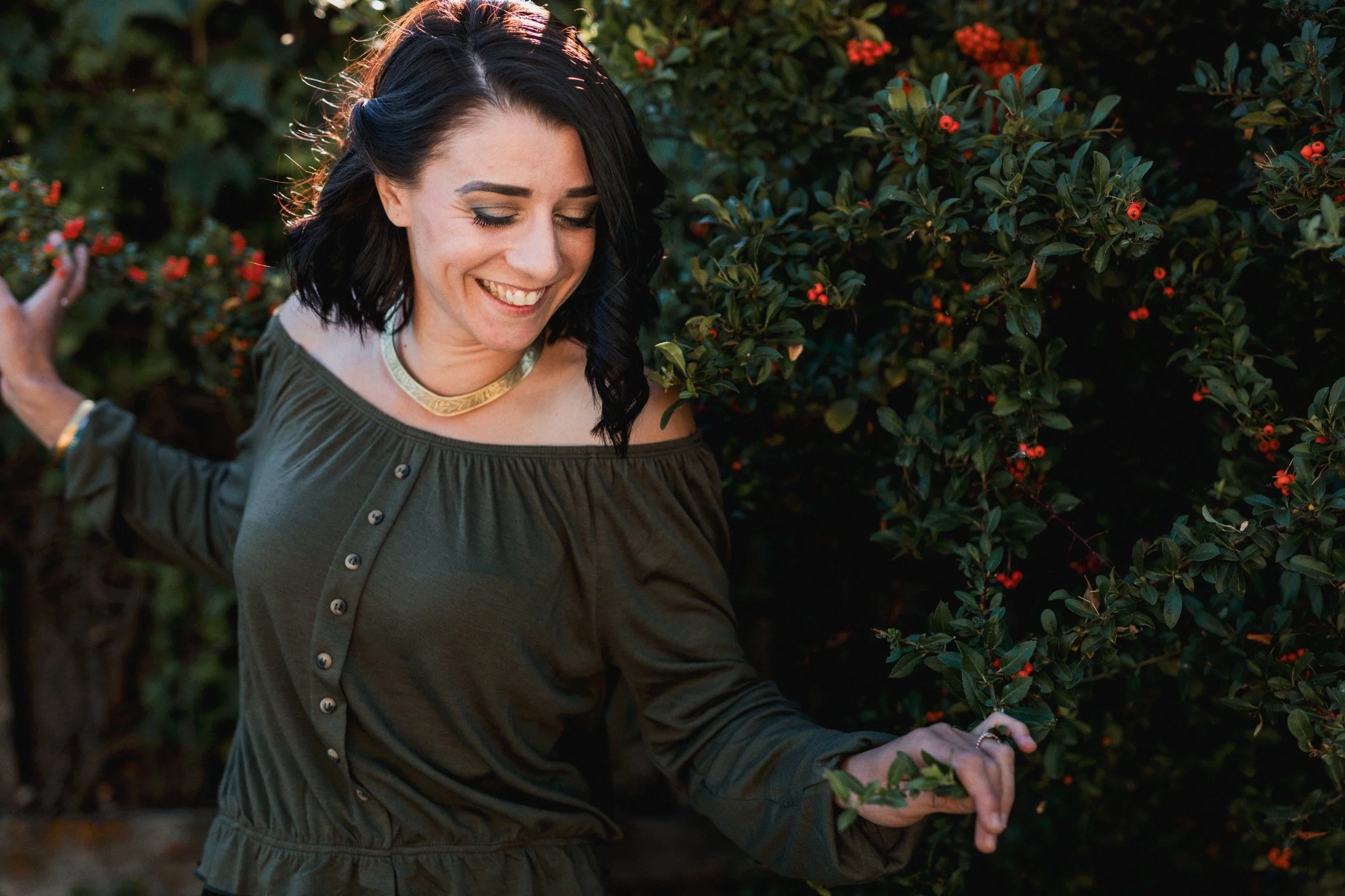 A woman with black hair smiling and looking down, touching plants with red berries on bushes behind her, outdoors in a garden or park.