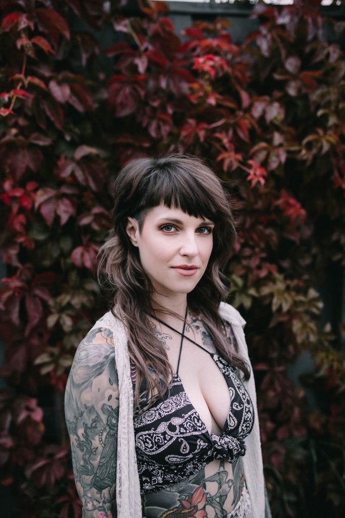 A woman with tattoos on her arms and chest, wearing a black and white patterned top and a light cardigan, standing outdoors in front of a bush with dark red leaves.