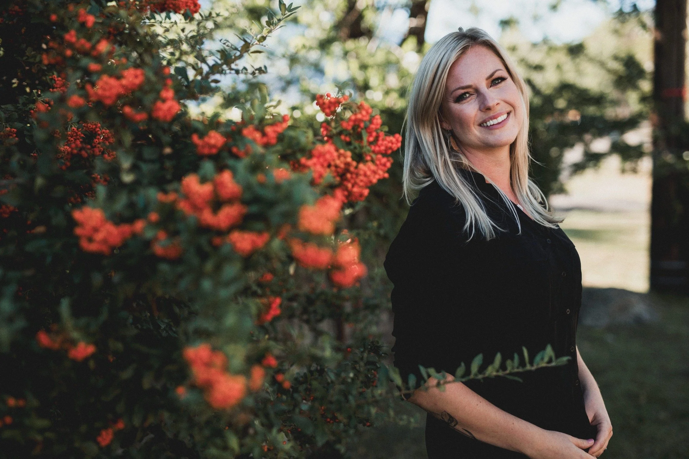 A smiling blonde woman standing outdoors near flowering bushes with red flowers, wearing a black shirt, with trees and blurred background in the distance.
