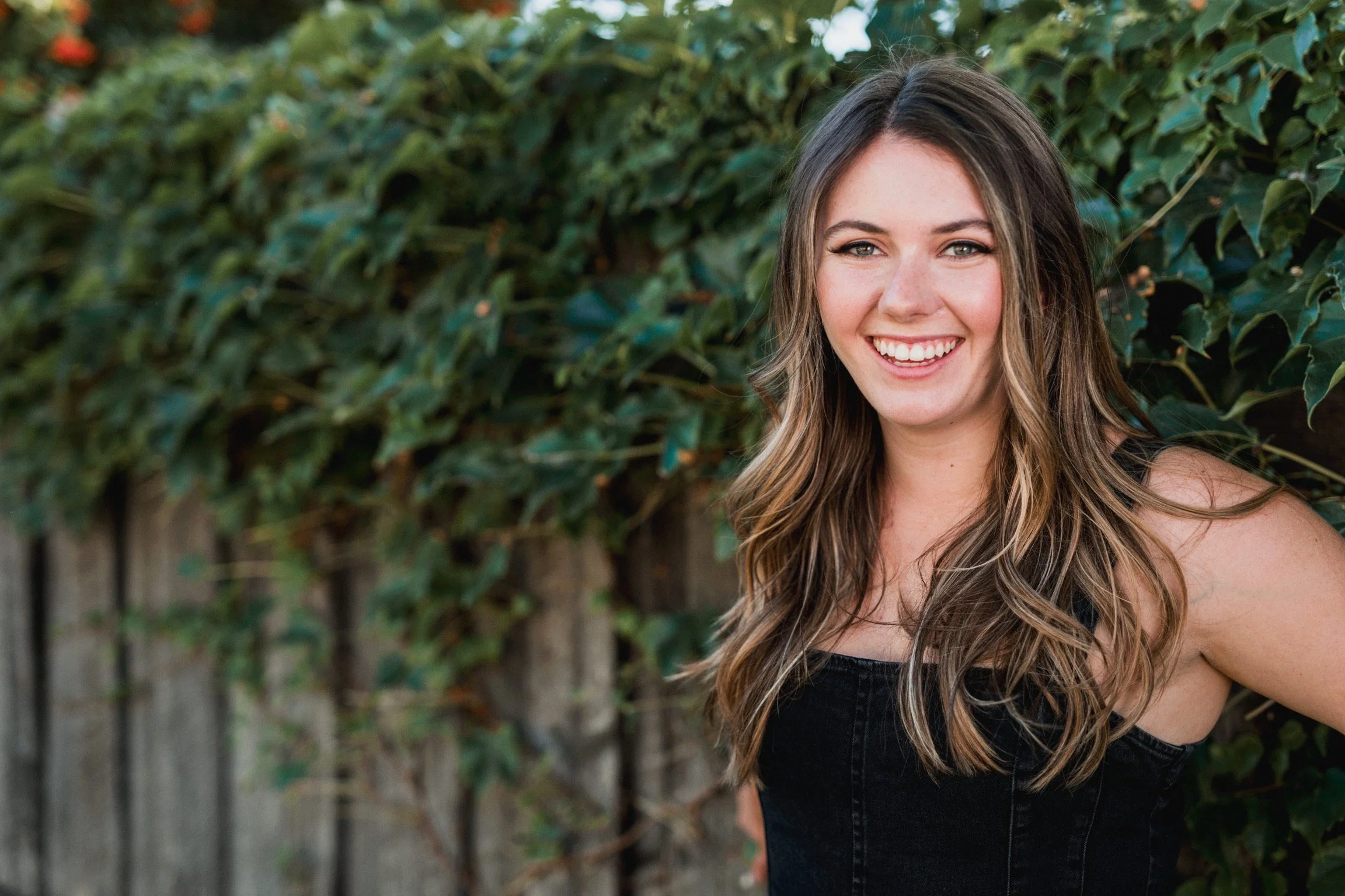 A young woman with long, wavy, brown hair smiling outdoors in front of a green hedge and wooden fence.