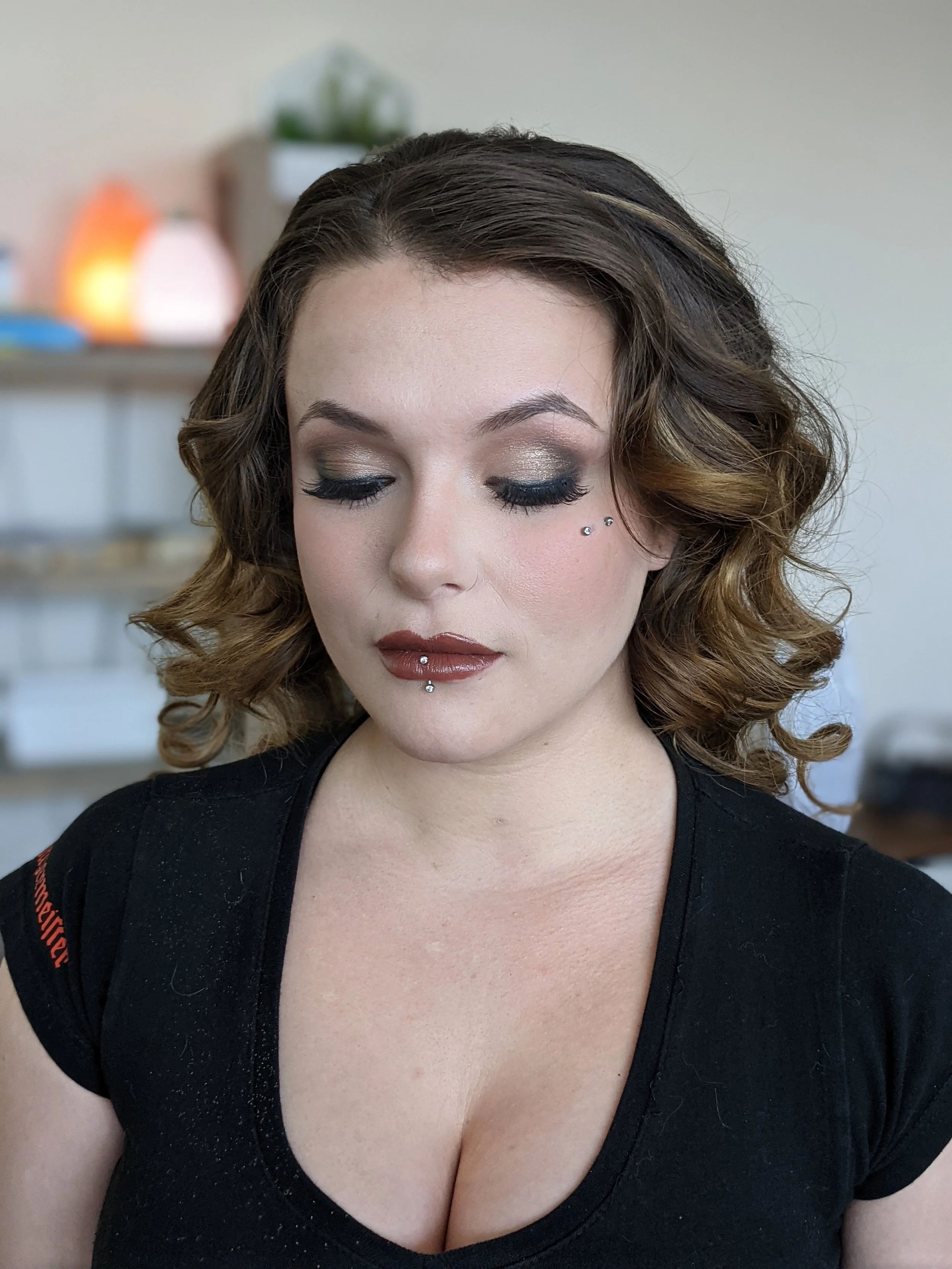A woman with wavy brown hair, closed eyes, wearing metallic eyeshadow, false eyelashes, facial piercings on her nose and lower lip, and a black top, in a room with blurred background.