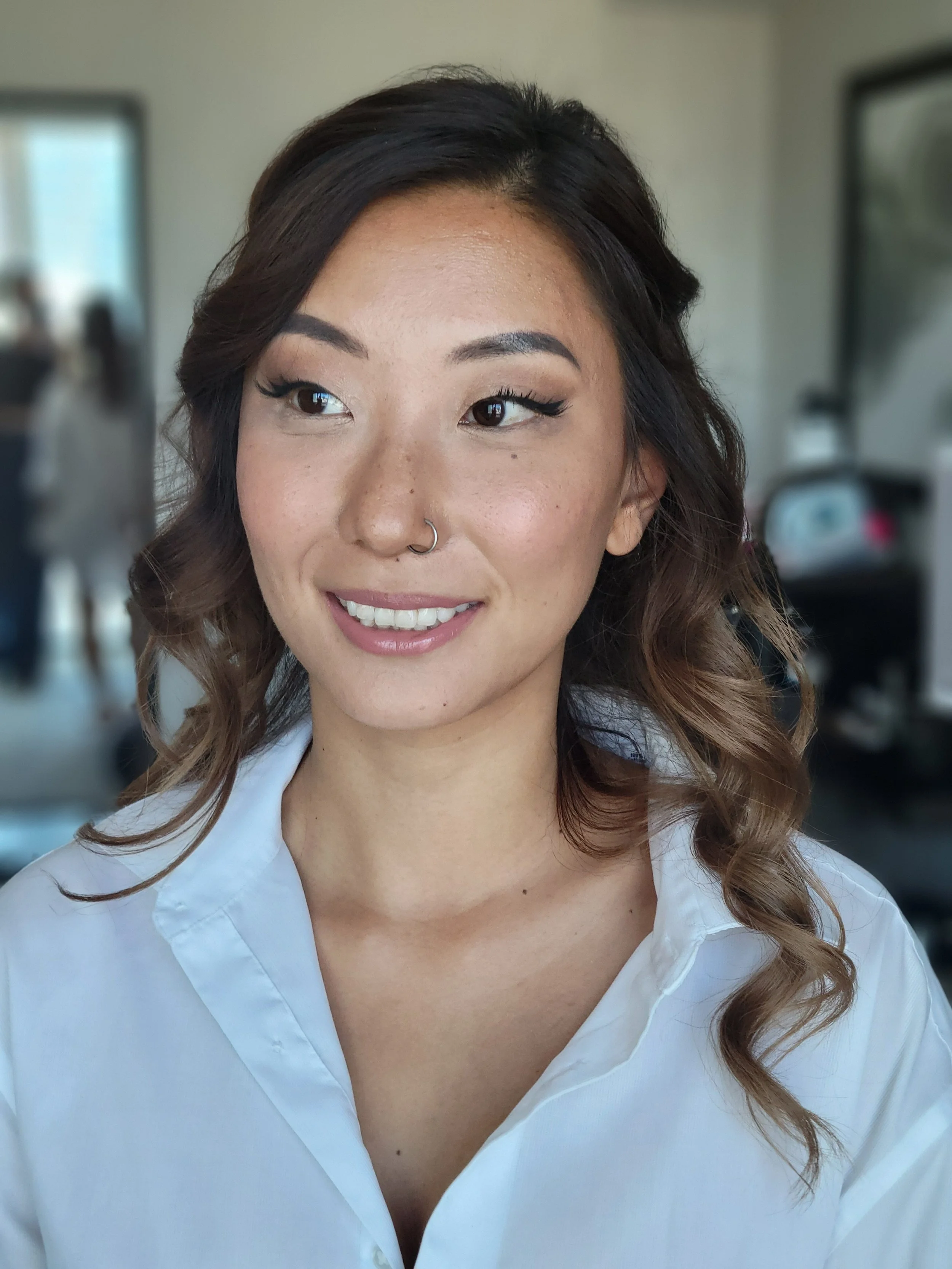 Close-up of a smiling woman with wavy hair, wearing a white shirt, in an indoor setting.