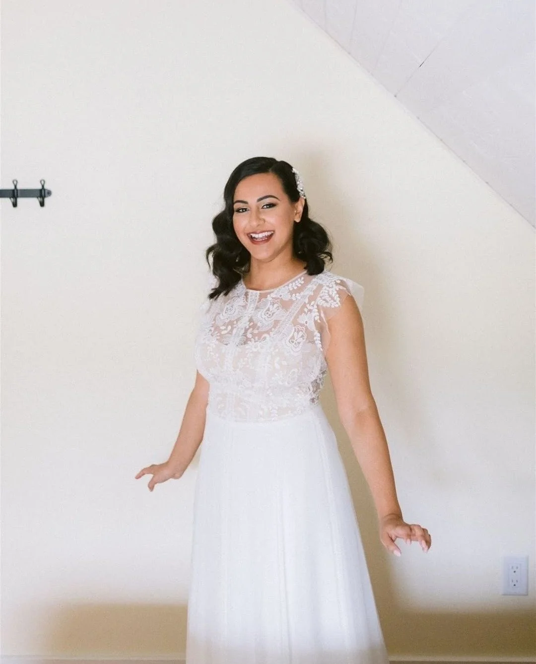 A smiling woman in a white lace wedding dress standing indoors against a plain wall with a sloped ceiling.