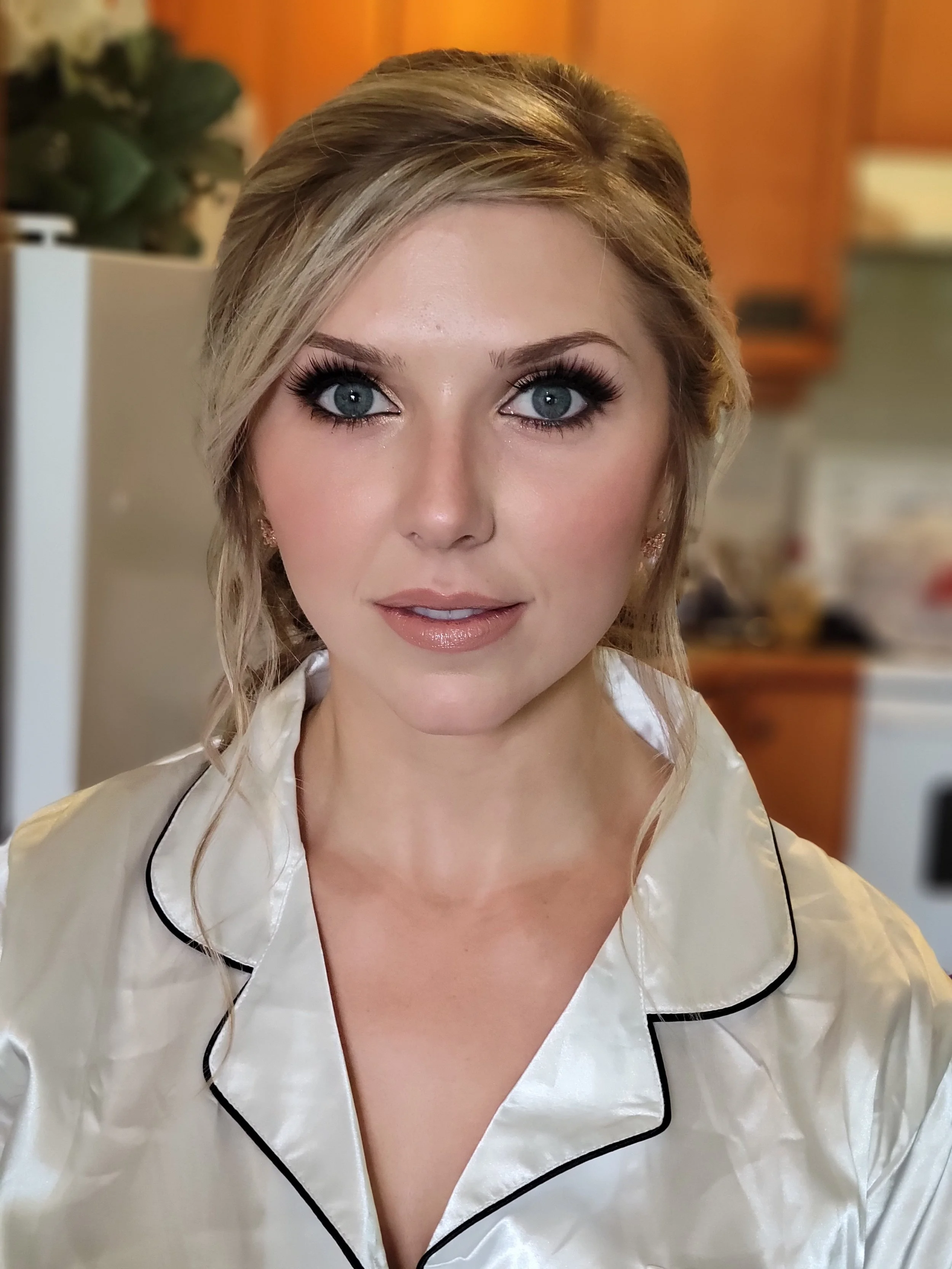 A woman with blonde hair and blue eyes wearing a satin white silk pajama top with black piping, sitting in a room with warm-colored walls and wooden cabinets.