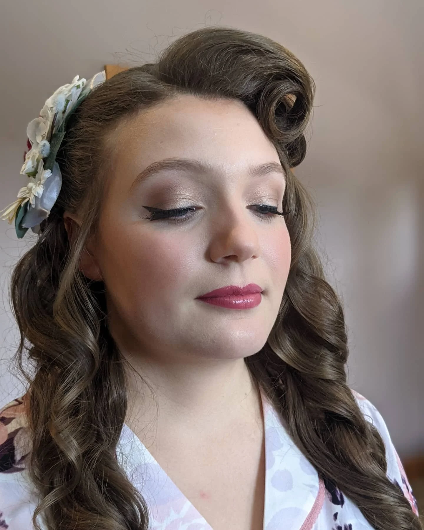 A young woman with vintage style hair curled in loose ringlets, wearing a floral headband, with makeup including winged eyeliner, shimmery eyeshadow, and pink lipstick, dressed in a light-colored blouse with a floral pattern.