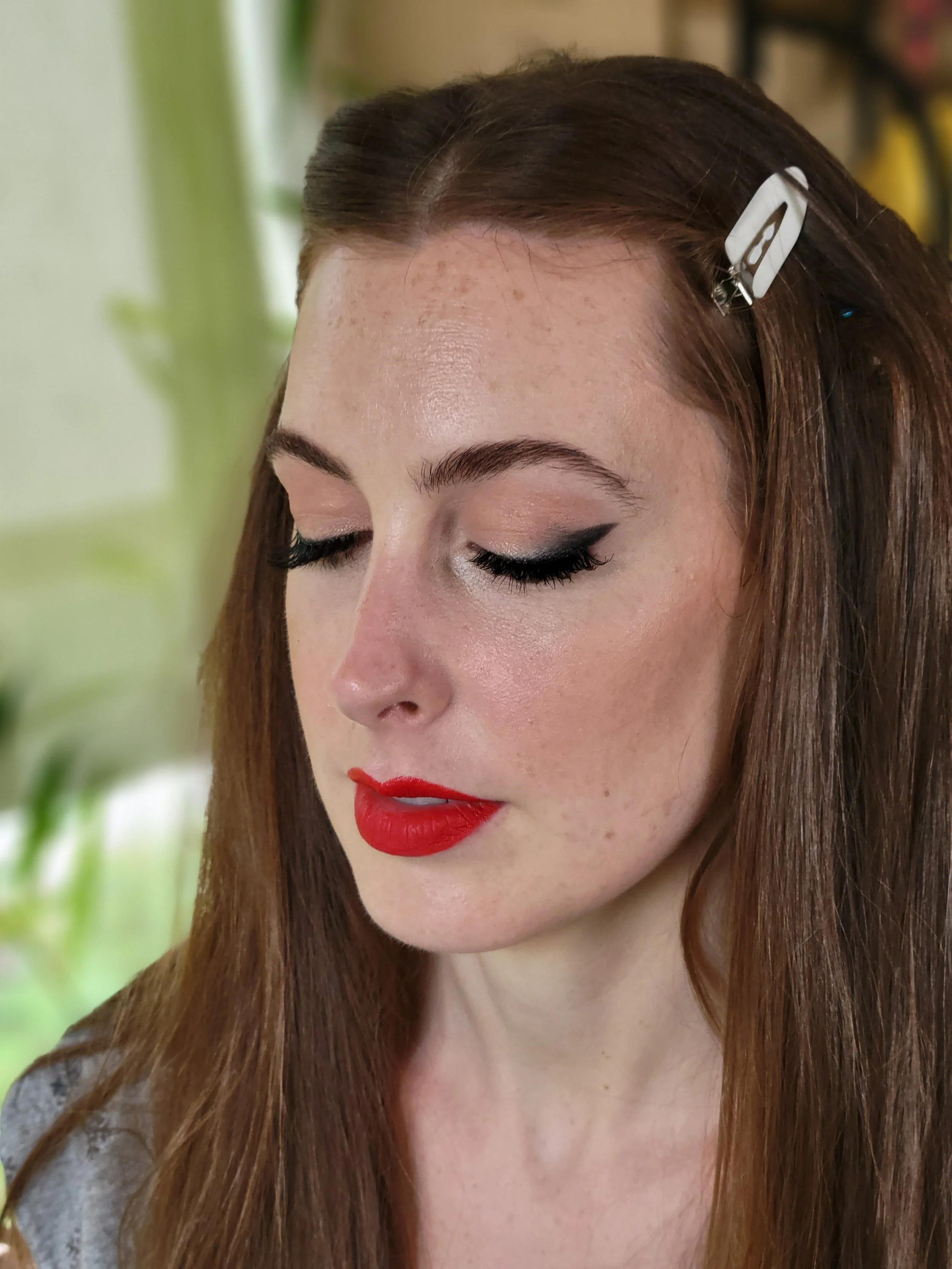 A close-up of a woman with long auburn hair, closed eyes with black winged eyeliner, red lipstick, and a white hair clip in her hair.