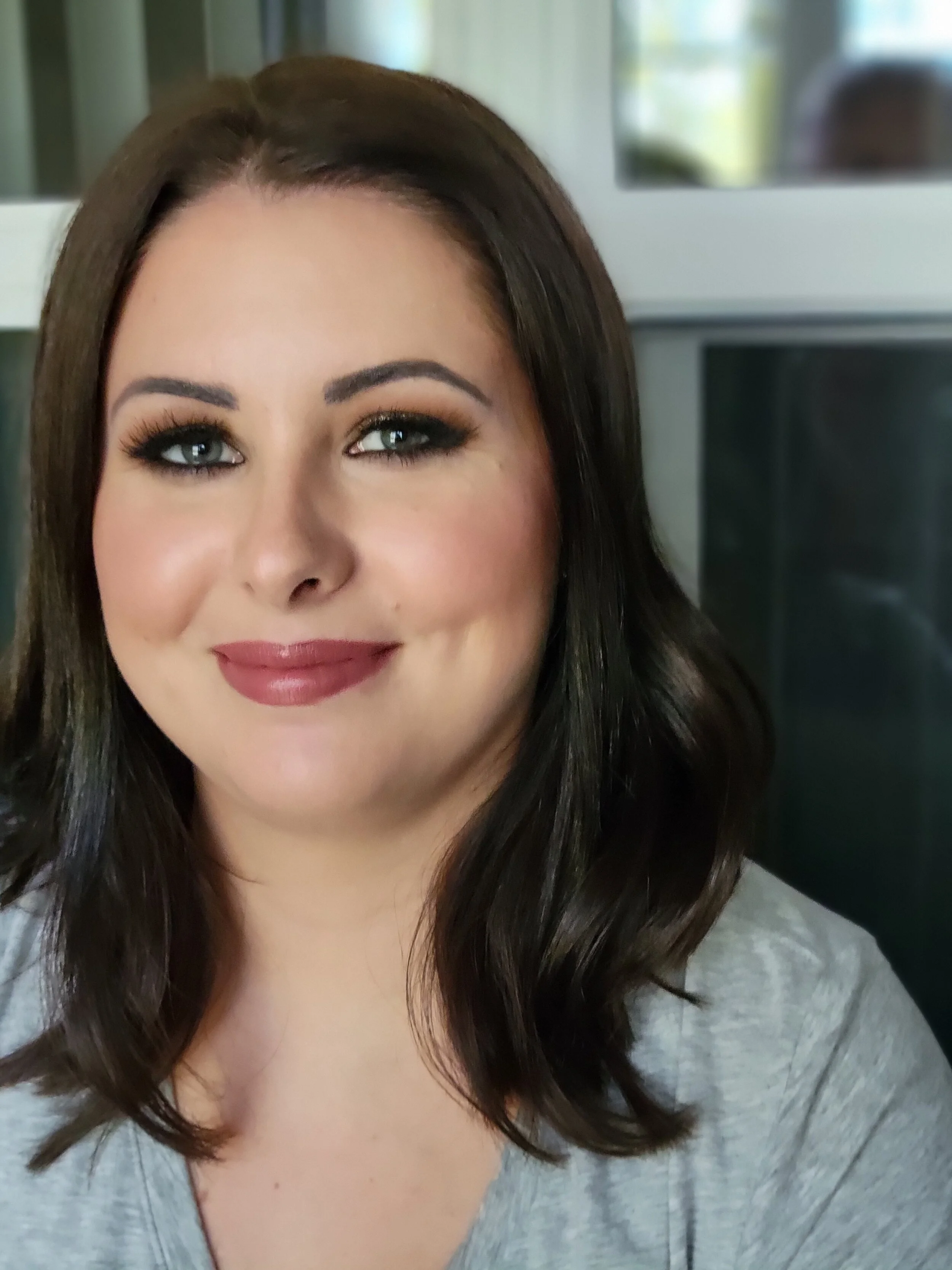 Close-up of a woman's face with makeup, dark hair, and wearing a gray top, smiling softly. Background includes a window with a view outside.