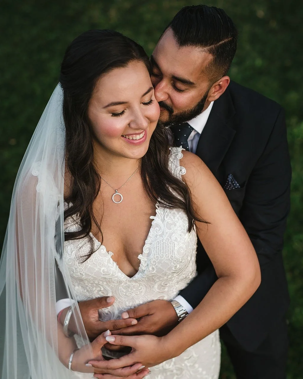 A newlywed couple embraces outdoors, smiling and sharing a tender moment with a lush green background.