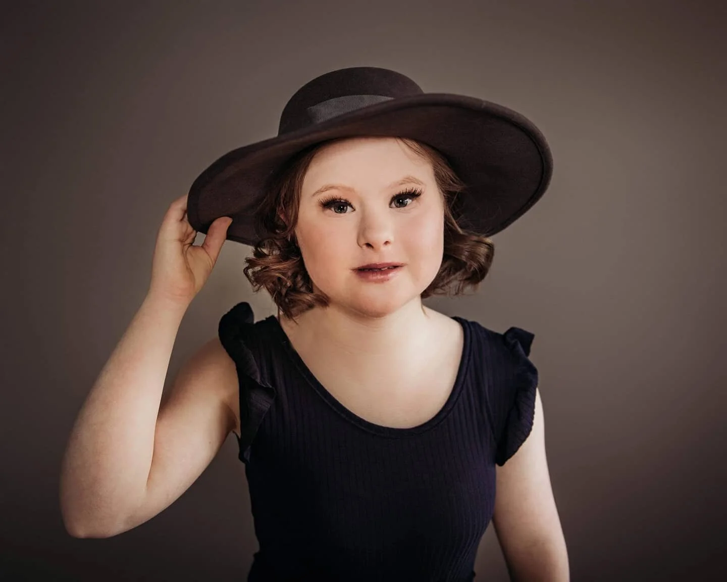 Portrait of a woman with curly hair wearing a black sleeveless top and a large black hat, holding the brim of her hat with her right hand, against a plain background.