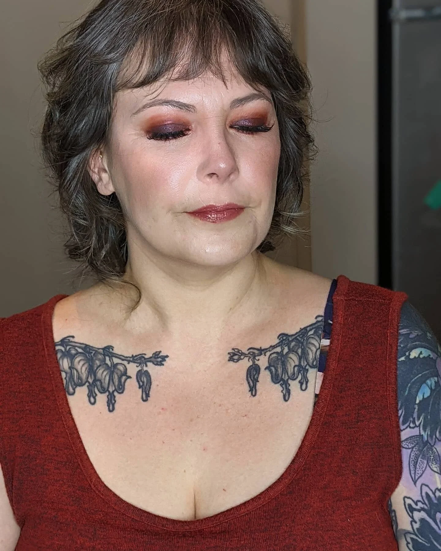 A woman with short brown hair wearing makeup, a red top, and displaying tattoos on her chest and arms.