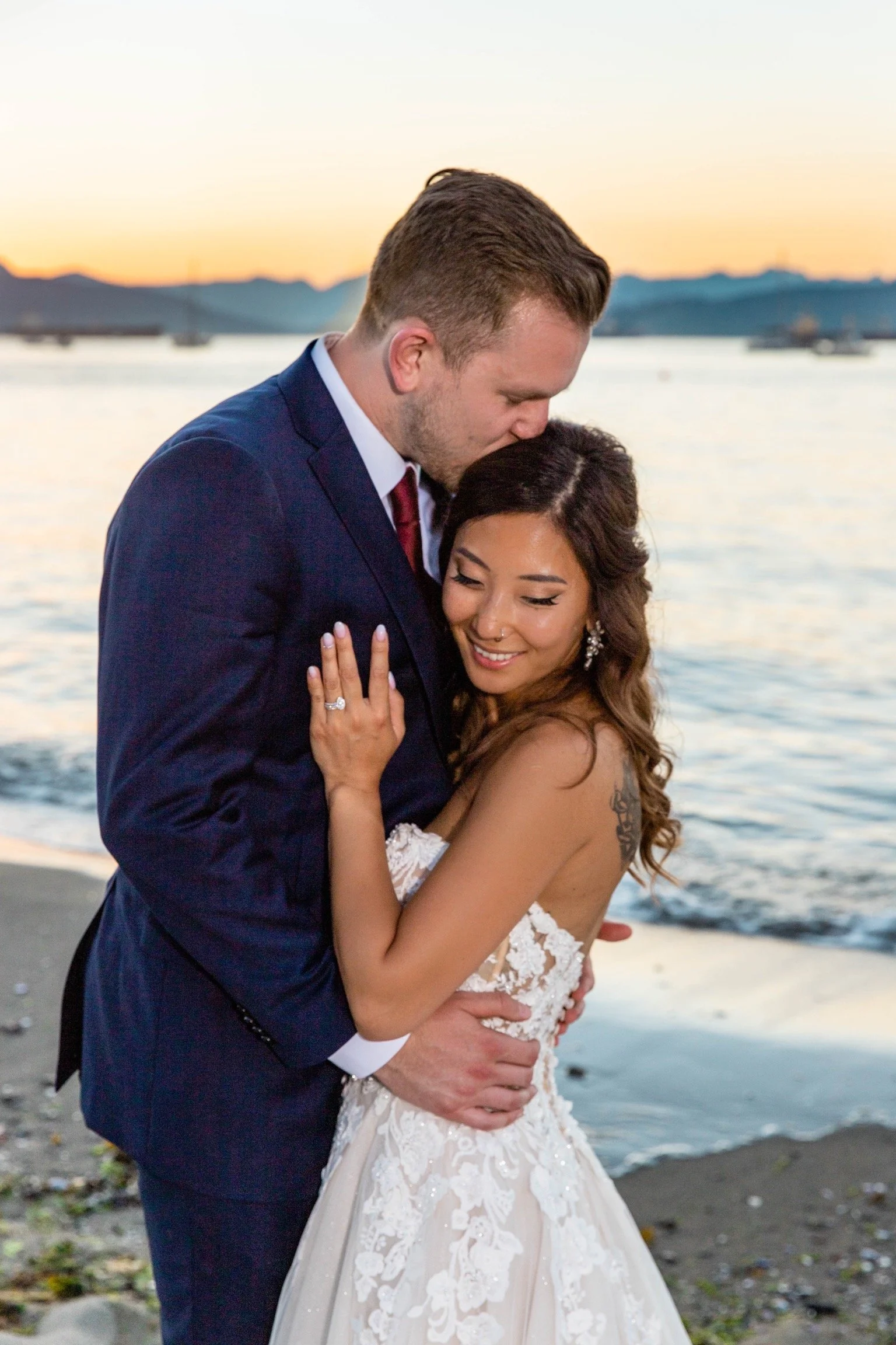 A newlywed couple embracing on a beach at sunset, with mountains and boats in the background.