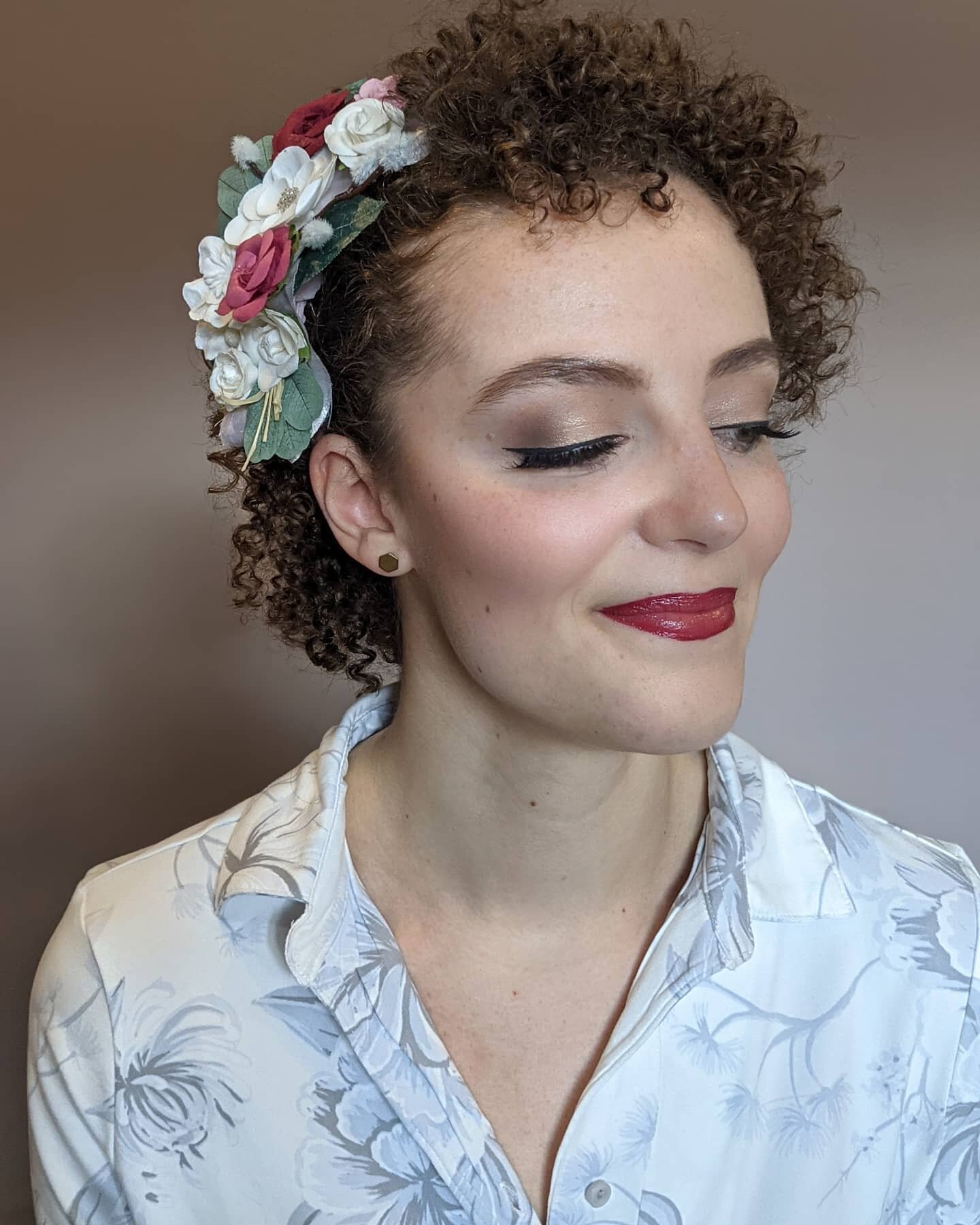 A woman with short, curly hair wearing a floral headband, makeup, and a white shirt with a subtle floral pattern, smiling slightly with eyes closed.