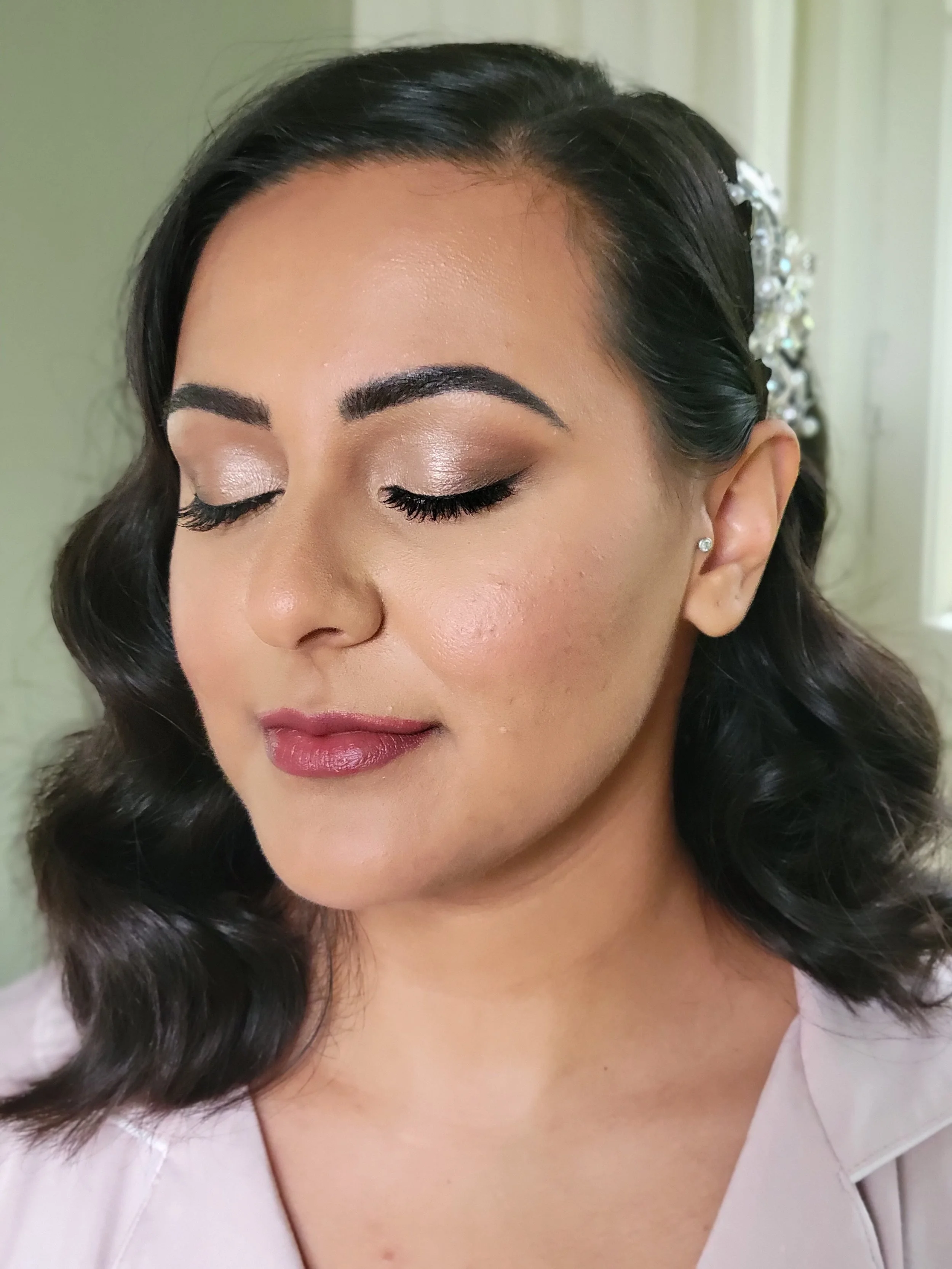A woman with makeup, closed eyes, dark wavy hair, and a pearl hair accessory, wearing a light-colored top.