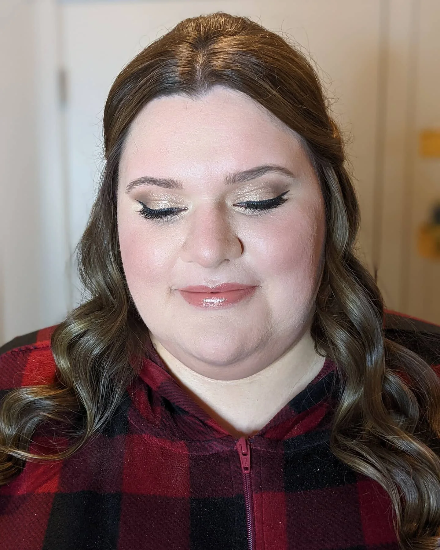 Woman with brown hair styled in loose curls, wearing gold eyeshadow, black eyeliner, and nude lipstick, dressed in a red and black plaid jacket with a zipper.