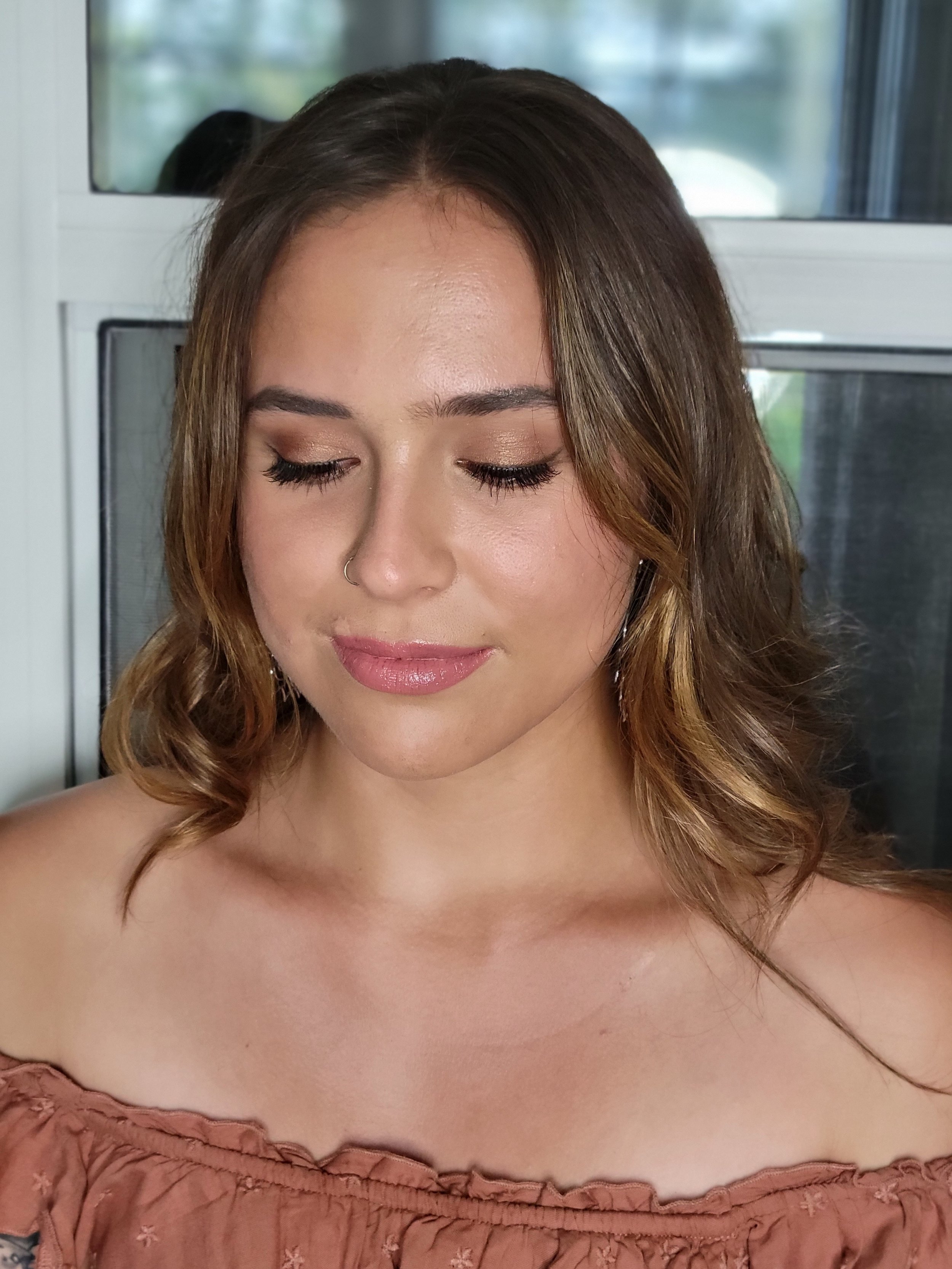 A woman with wavy brown hair and closed eyes, wearing makeup, and an off-shoulder, ruffled, peach-colored top.