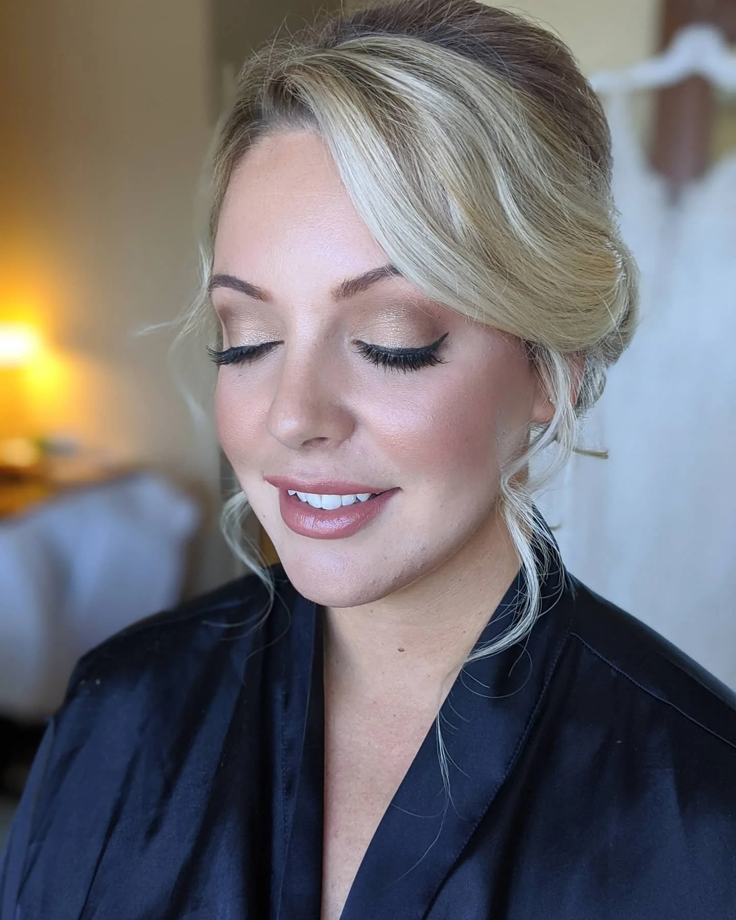 A woman with blonde hair styled in loose curls, wearing makeup with nude lipstick and eye makeup, dressed in black, smiling softly with closed eyes in a warmly lit room.