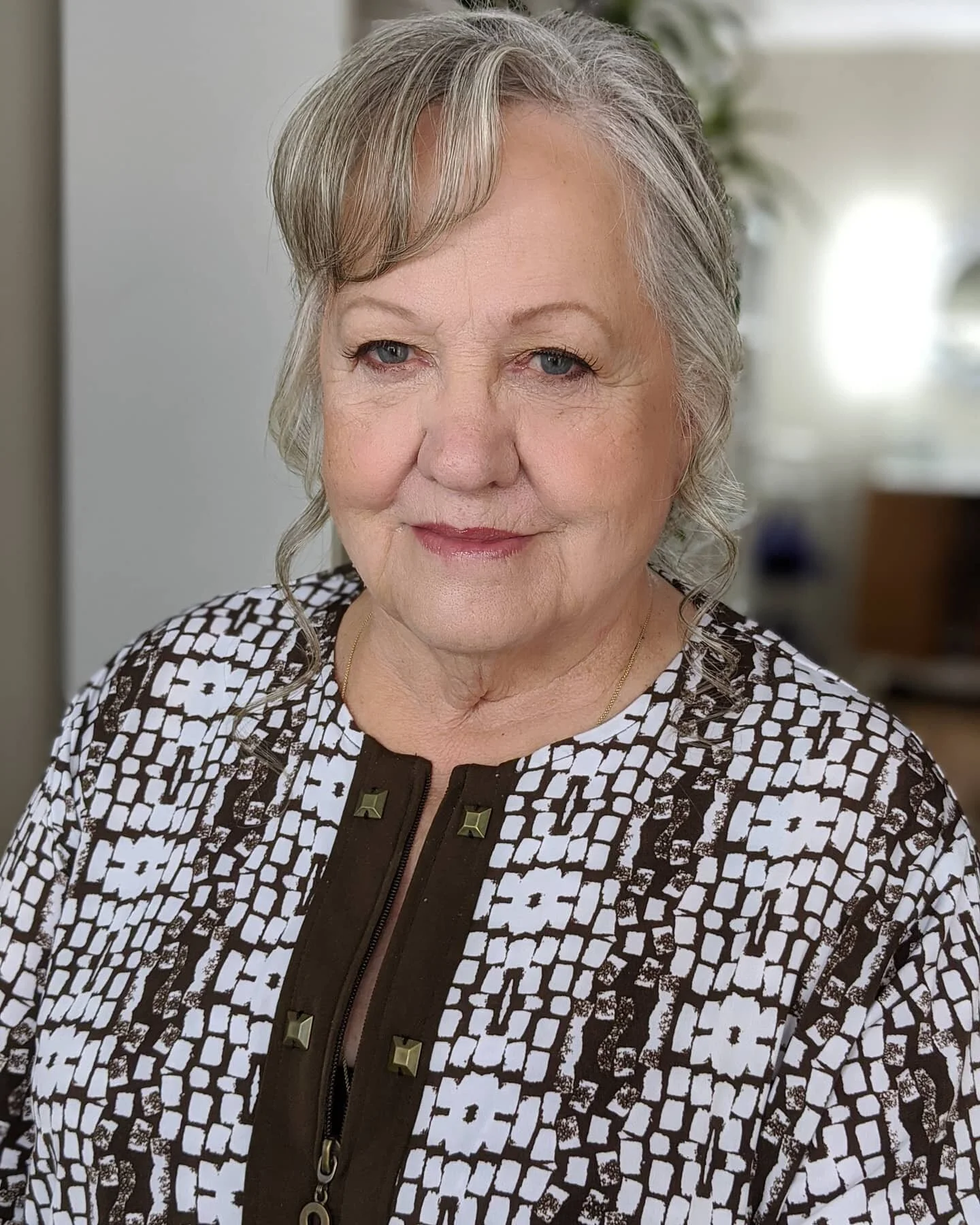 A smiling elderly woman with short, gray hair, wearing a patterned black and white top with a zipper and gold buttons, standing indoors with a blurred background.
