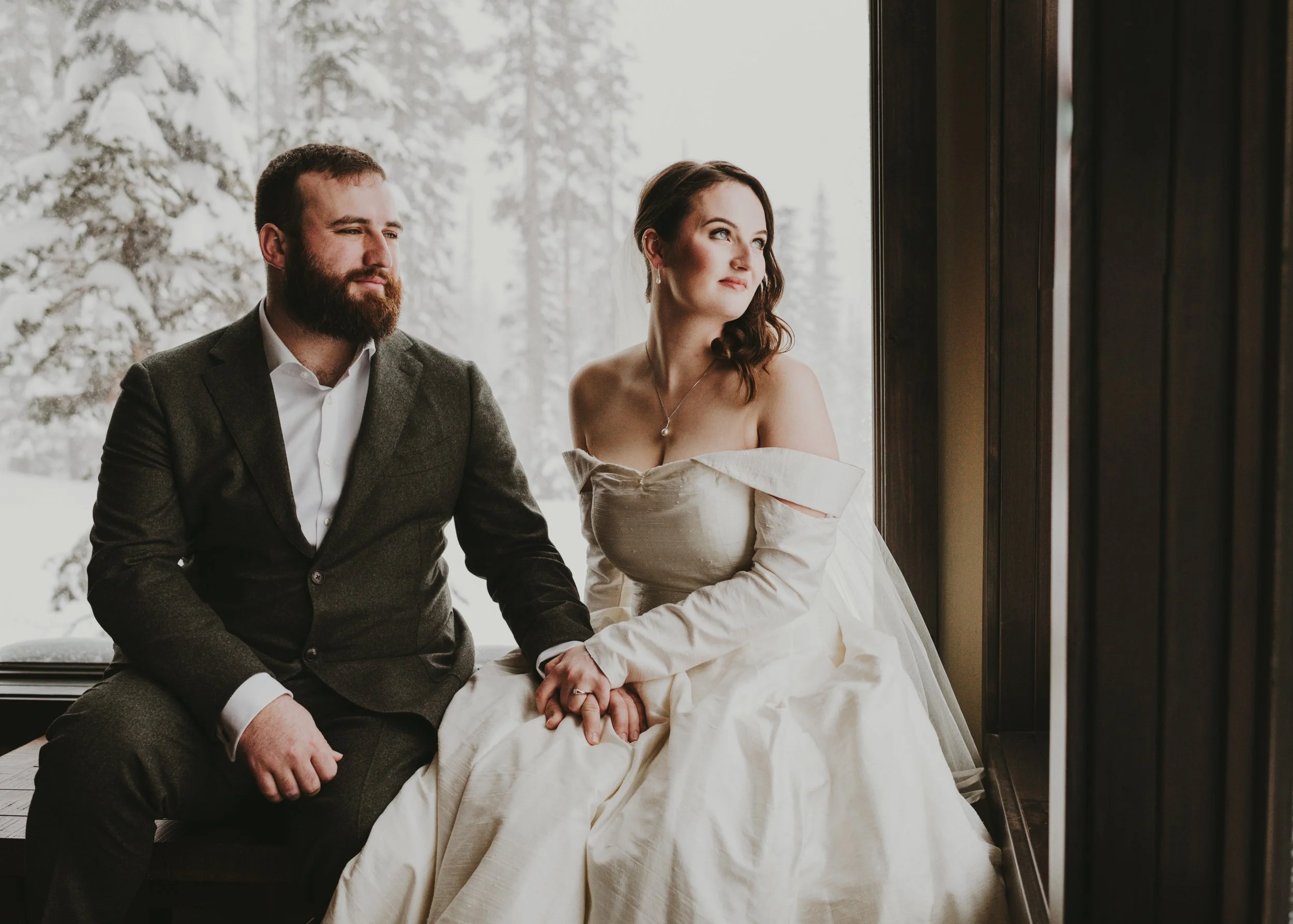 A man and woman sitting closely together indoors near a large window with a snowy outdoor landscape, the woman is wearing a white dress and the man is in a gray suit.
