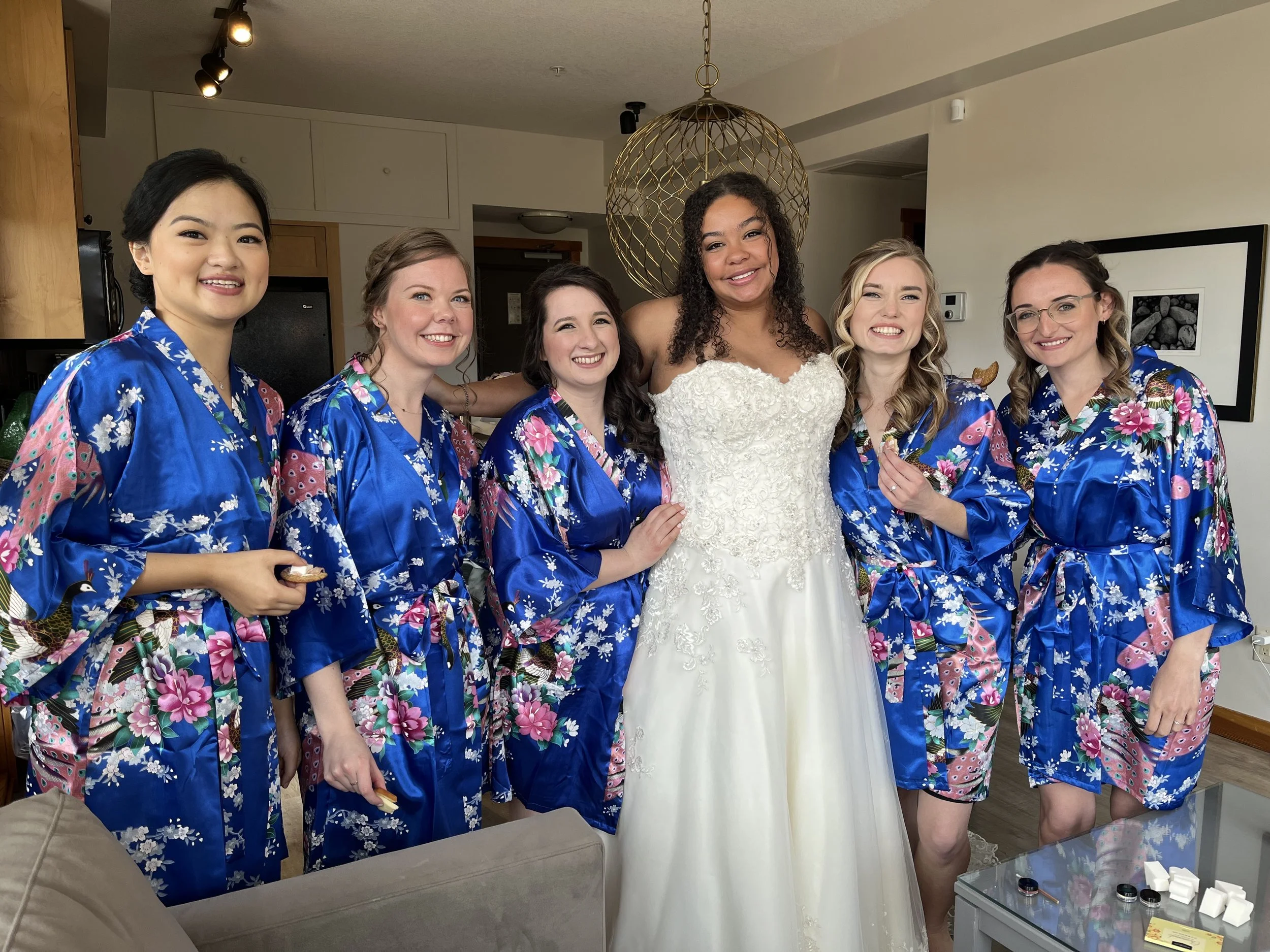 Group of women, one in a white wedding dress, and five others in matching blue floral robes, posing indoors during a wedding preparation.
