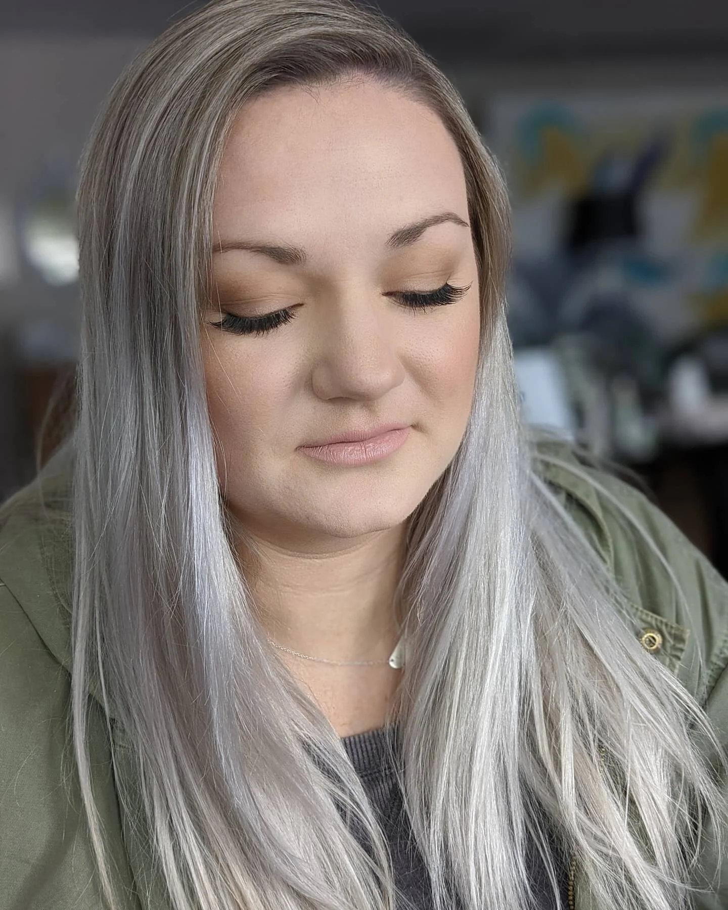 A woman with long, silver hair, closed eyes, wearing a green jacket, and a delicate necklace, in an indoor setting.