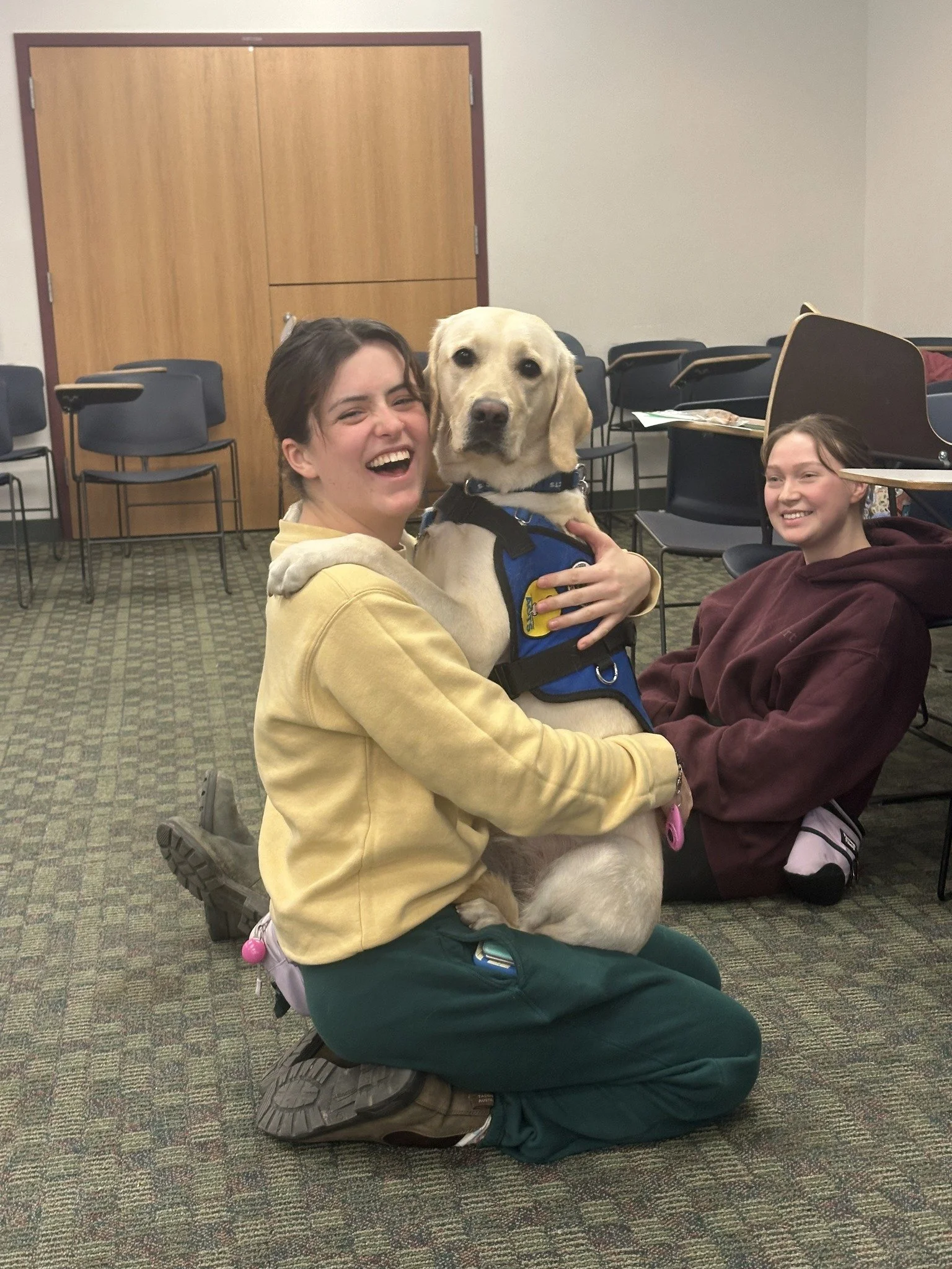 A woman wearing a yellow hoodie and green pants is kneeling on the floor, hugging a large yellow Labrador retriever with a service harness. She is smiling and laughing. Seated behind her is a young girl in a maroon hoodie, sitting on the floor, smiling. The room has chairs and tables stacked or pushed to the sides, with a wooden door in the background.