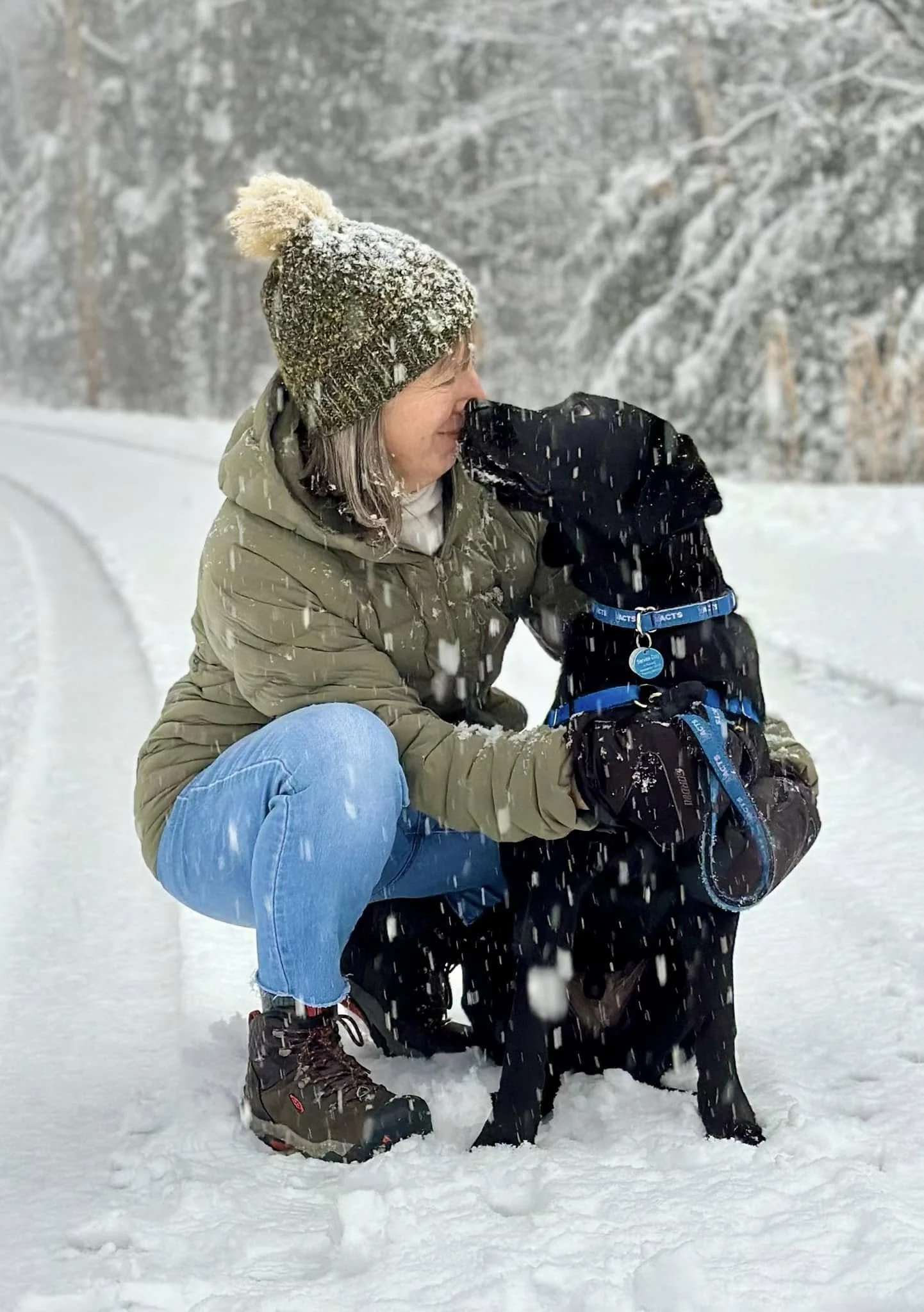 A woman in winter clothing squats in the snow, hugging her black Labrador Retriever dog during a snowfall in a snowy landscape.