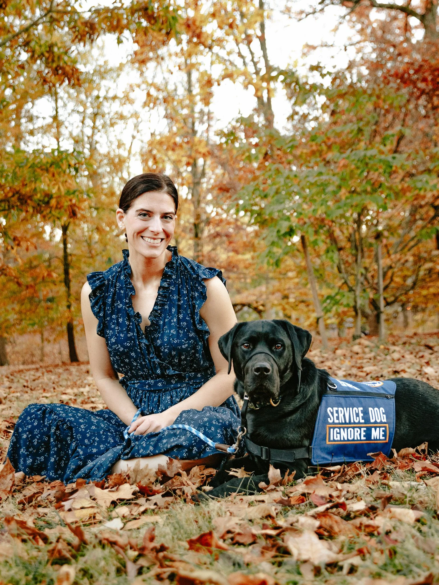 A woman in a blue dress sitting on the ground with a service dog, a black Labrador, in an autumn forest with fallen leaves and colorful trees.