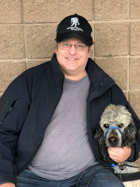 A man with glasses and a black cap sitting next to a dog with a harness, against a brick wall.