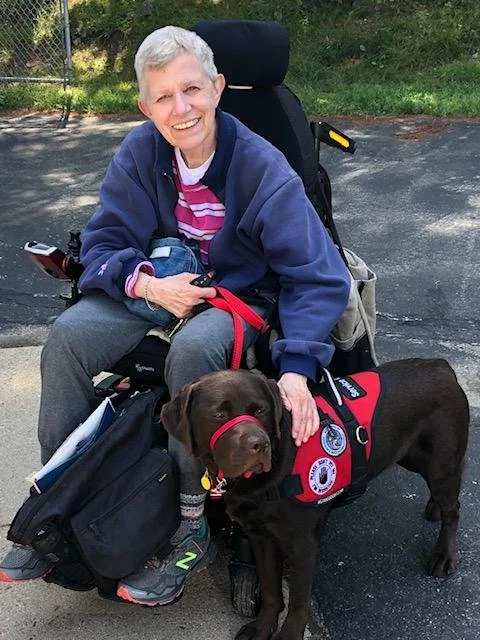An elderly woman sitting in a wheelchair outdoors with a chocolate Labrador dog wearing a red service vest, smiling.